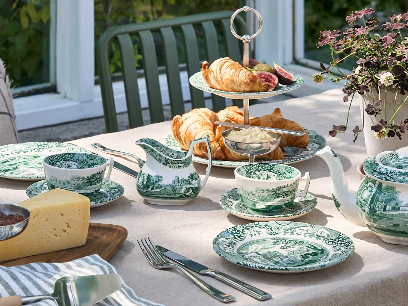An outdoor table set with green and white patterned dinnerware, croissants, cheese, and a teapot, in a garden setting.