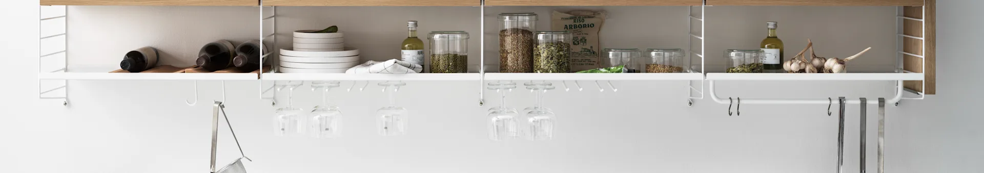 White kitchen shelving unit with wine bottles, plates, dry goods, olive oil, garlic, and hanging wine glasses and utensils.