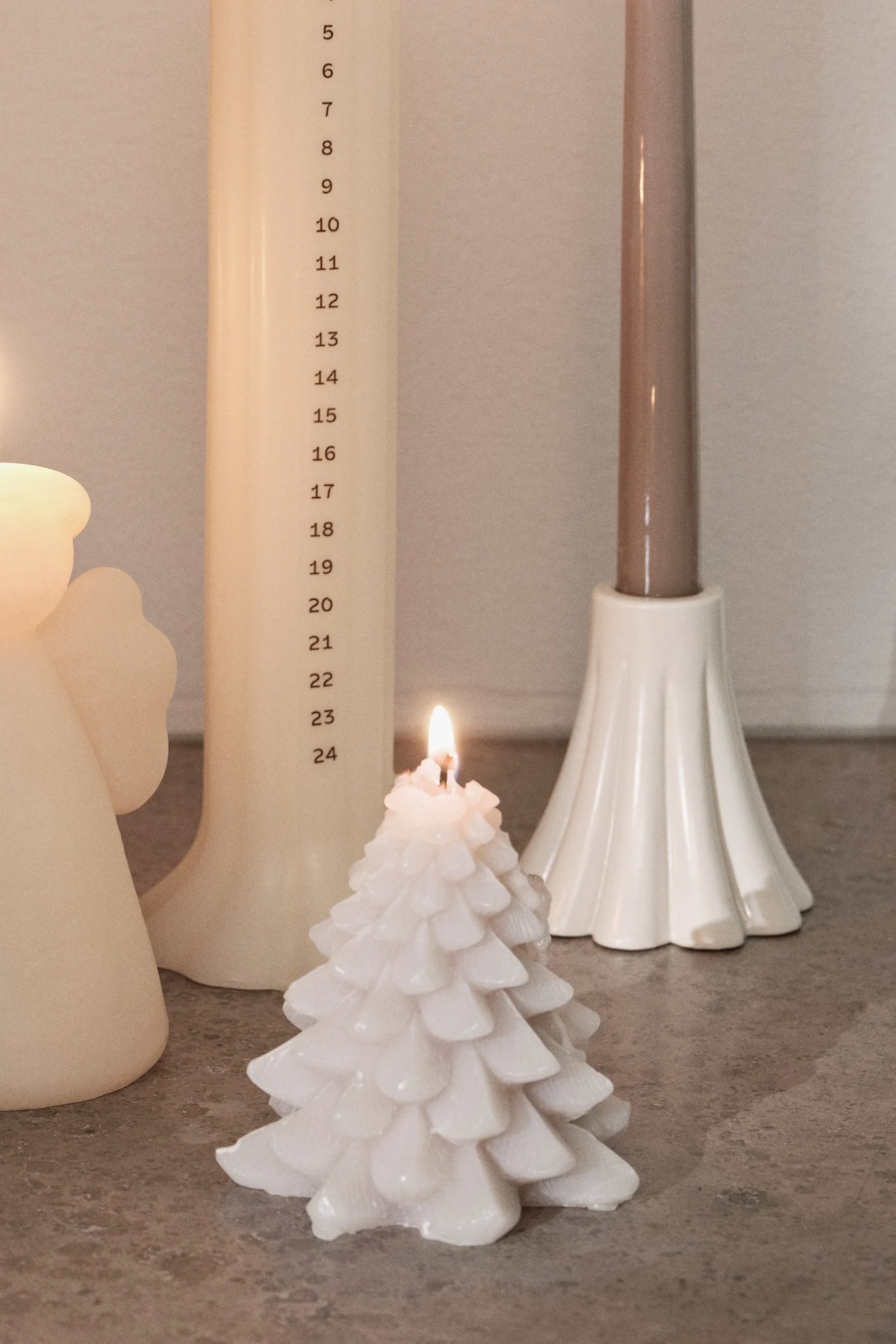 A lit white Christmas tree candle, an advent candle with numbers 5-24, and a brown candle in a white holder on a gray surface.
