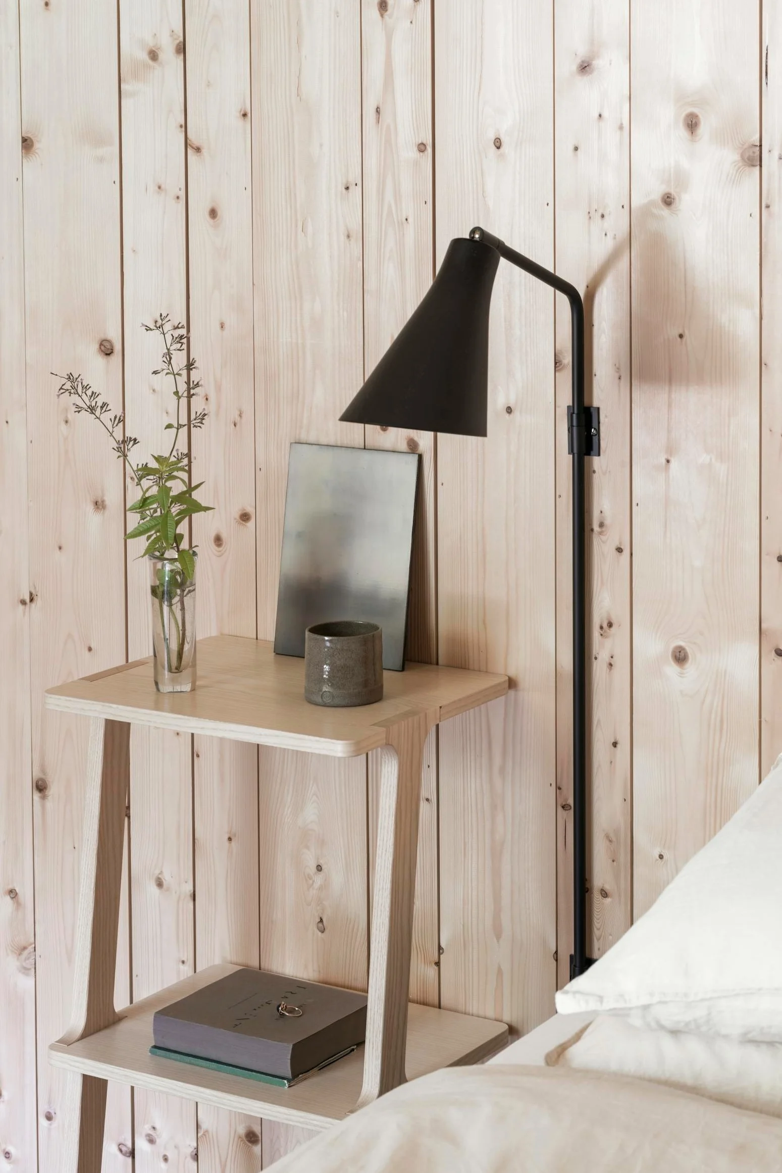 A minimalist light wood nightstand with a vase, mug, and book, next to a bed with white linen and a black wall lamp.