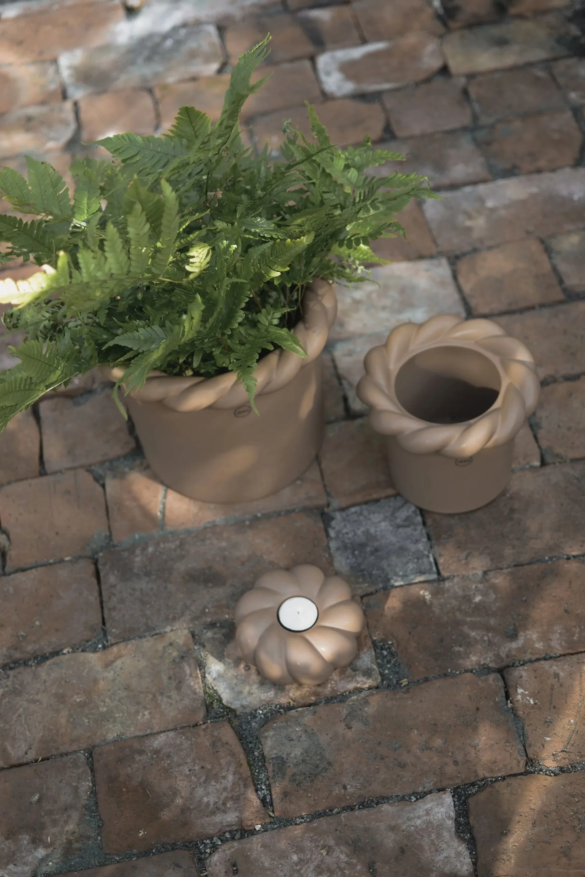 A potted fern, an empty pot, and a candle holder with a tea light sit on a brick patio, all pots feature a braided design.