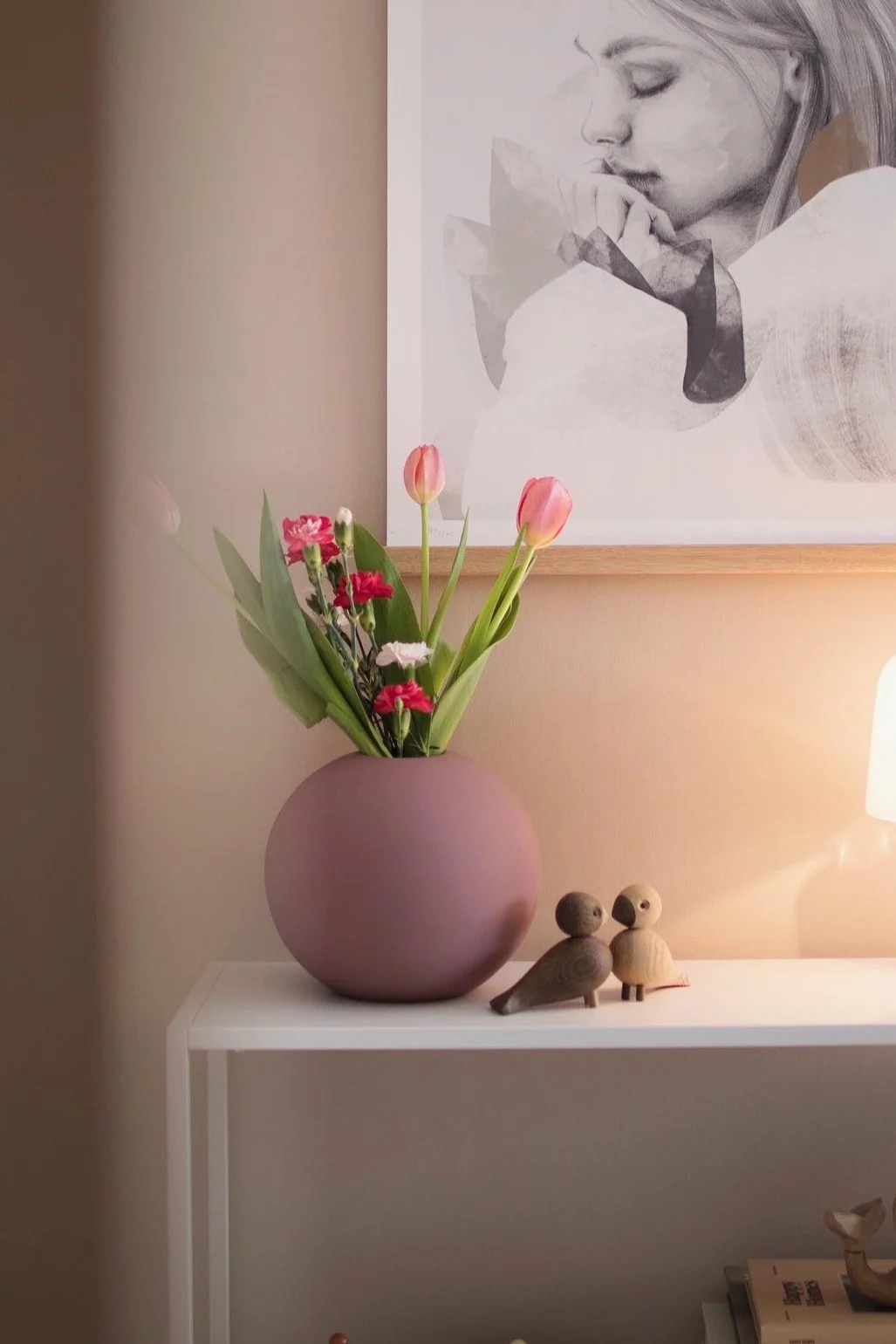Round purple vase with pink tulips and small flowers, two wooden birds, and a framed abstract portrait on a shelf.