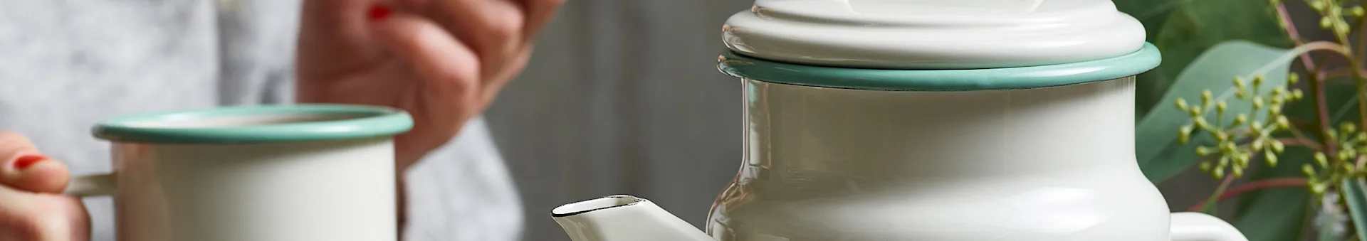 Close-up of off-white enamel mug and coffee pot with green rims, held by a person with red nail polish.
