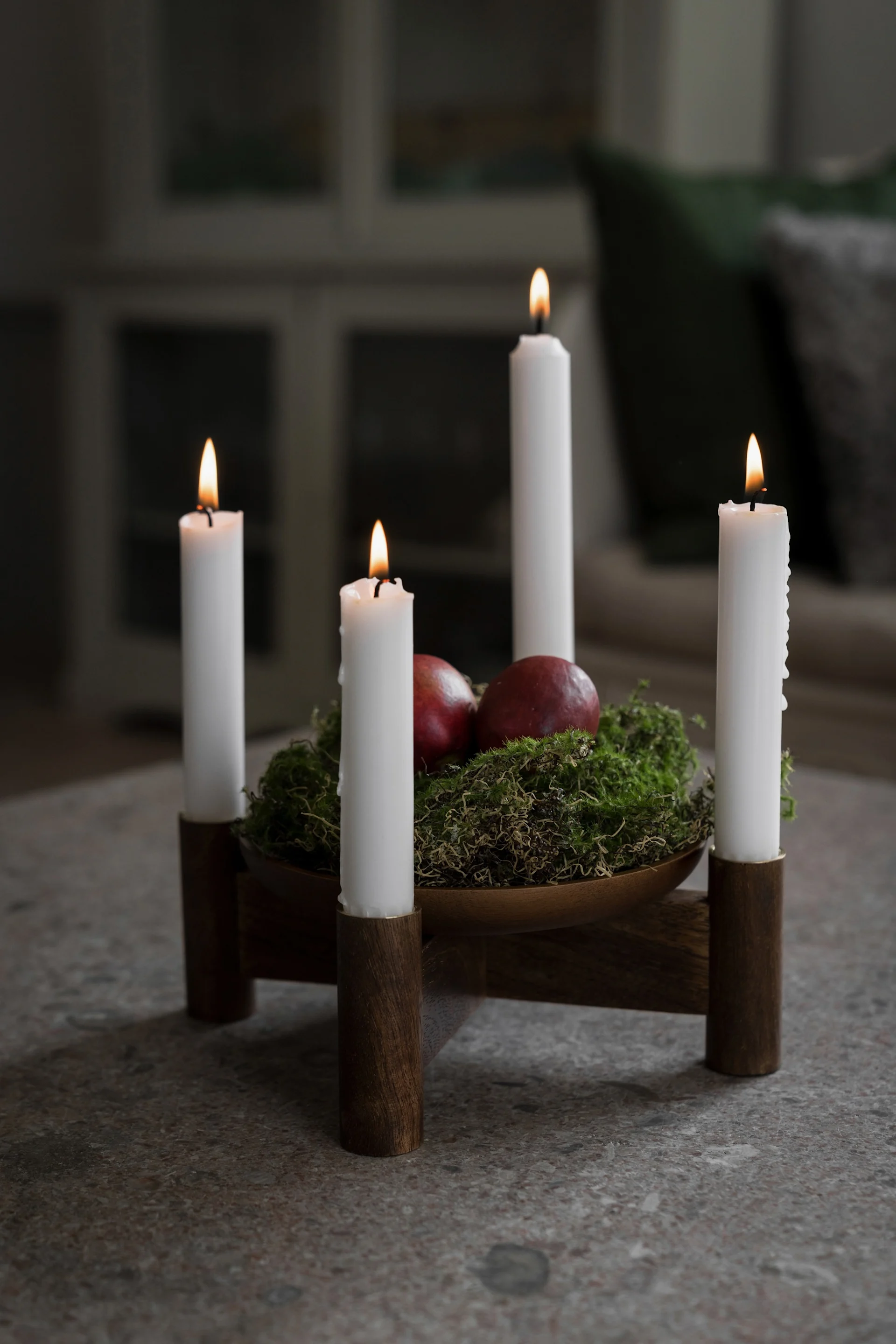 Wooden candle holder with four lit white candles, green moss, and two red pomegranates on a stone surface.