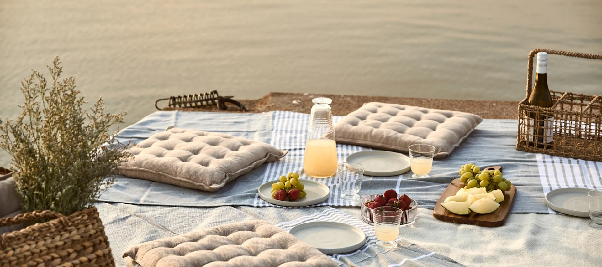 Picnic scene by the water with cushions, fruit, and drinks on a blanket.