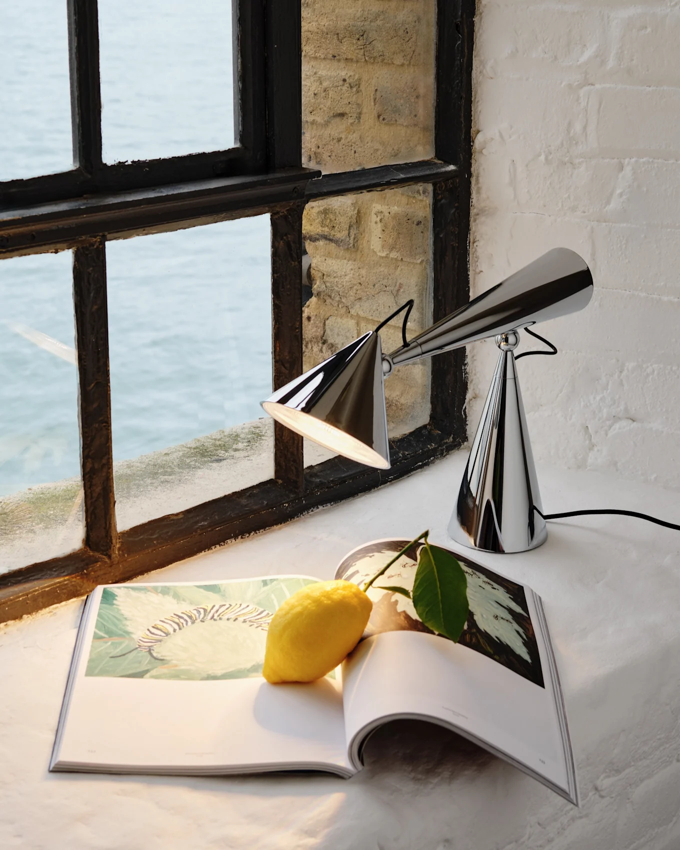 A chrome table lamp with two conical shades, an open book with a lemon, and a window overlooking water.