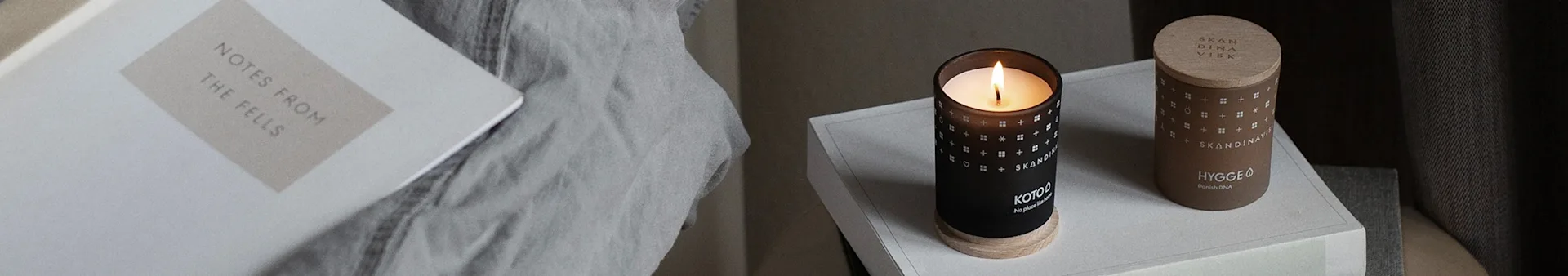 A book titled 'NOTES FROM THE FELLS' on a grey blanket next to two Scandinavian-style candles, one black and burning, one brown with a wooden lid.