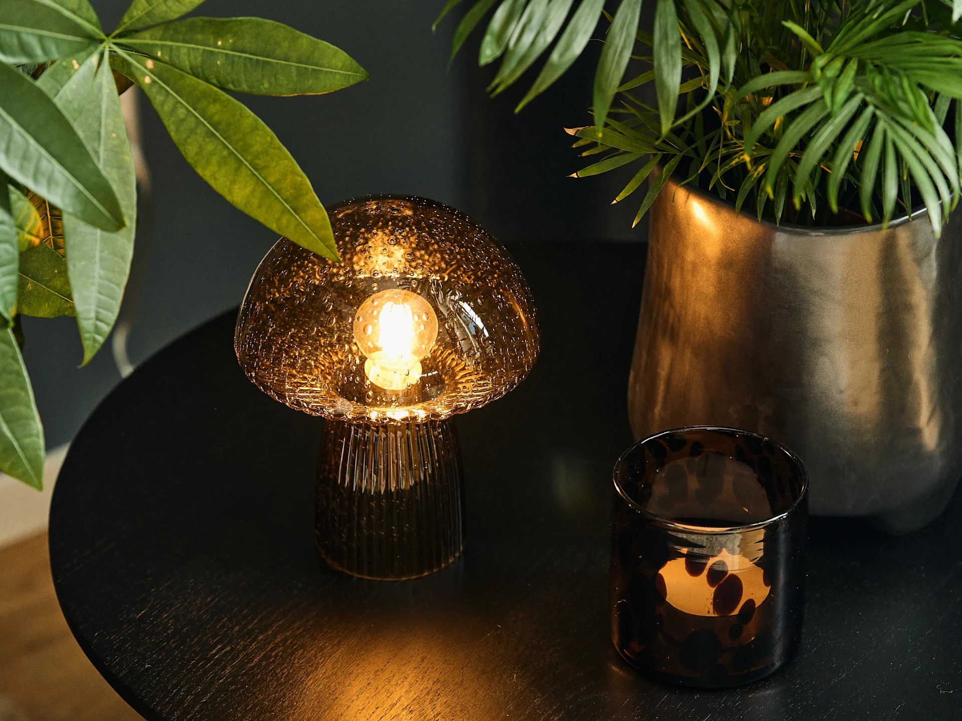 Mushroom-shaped glass table lamp and spotted glass candle holder on a dark table, flanked by green plants.