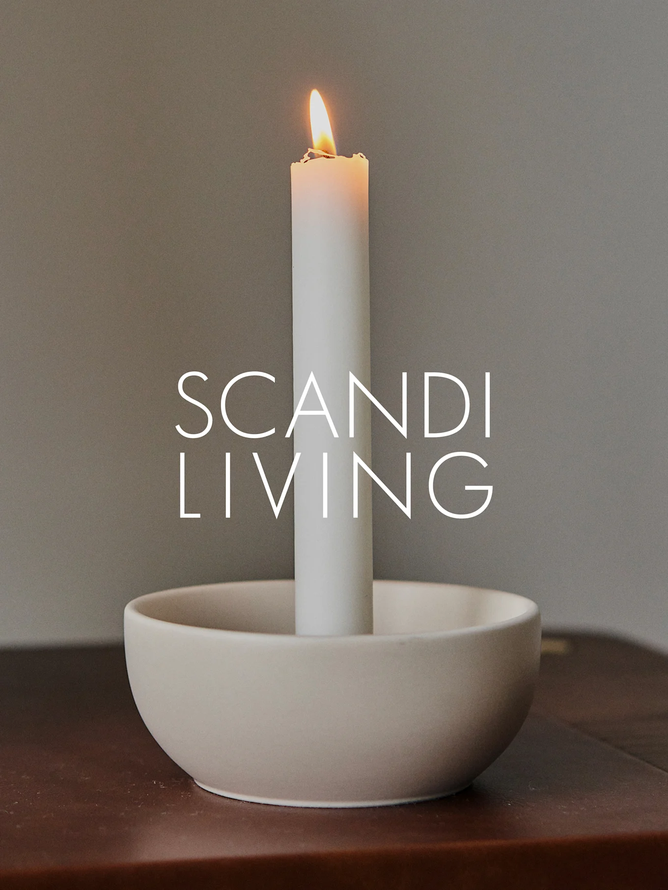 A lit white pillar candle in a light beige bowl-shaped holder on a dark wooden surface, with 'SCANDI LIVING' text overlaid.