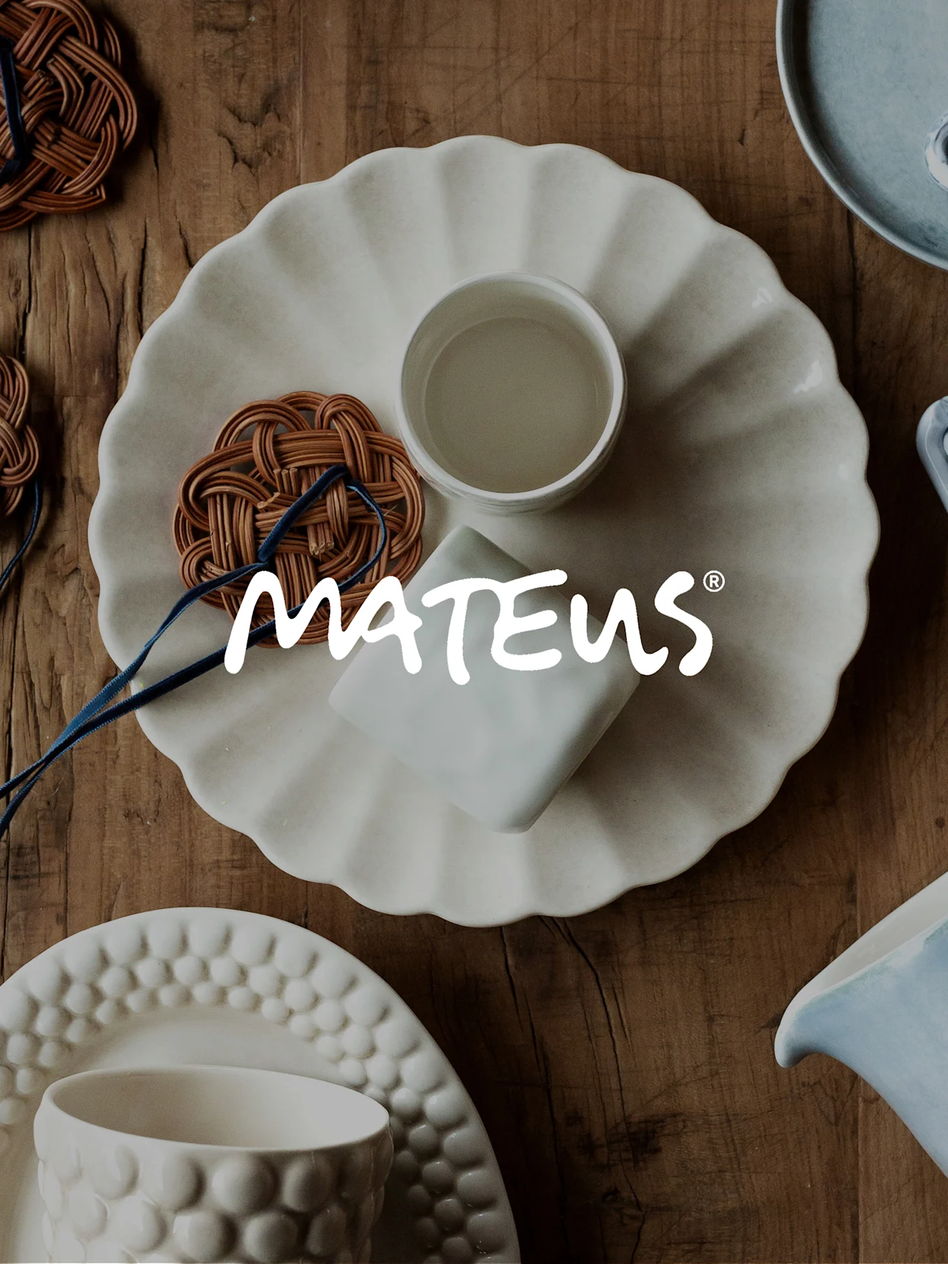 MATEUS ceramic tableware and woven coasters on a wooden table, featuring a ruffled plate, cup, and pitchers.