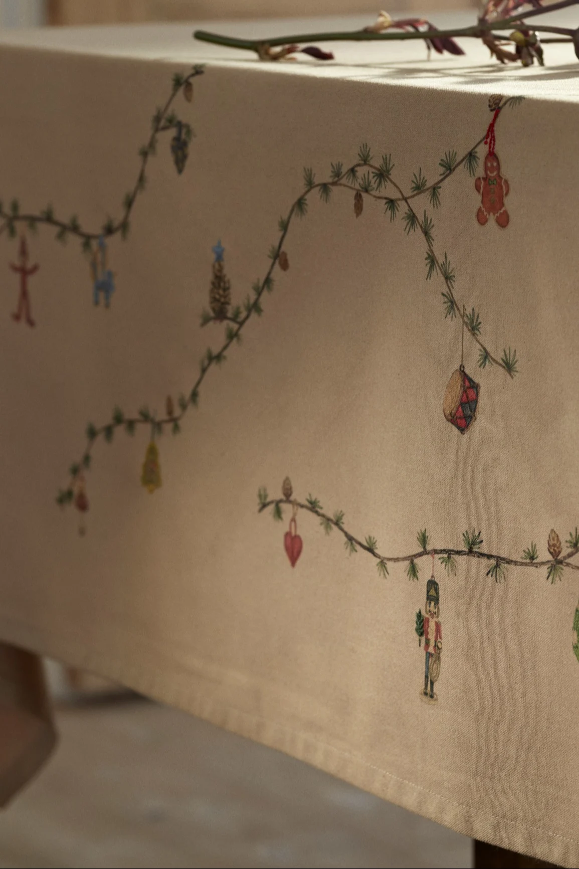 Light brown tablecloth decorated with painted Christmas garland featuring a gingerbread man, nutcracker, drum, and other ornaments.