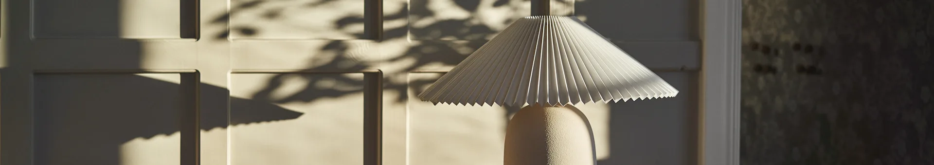 A table lamp with a cream pleated shade and a textured base, casting shadows on a cream panelled wall from sunlight.