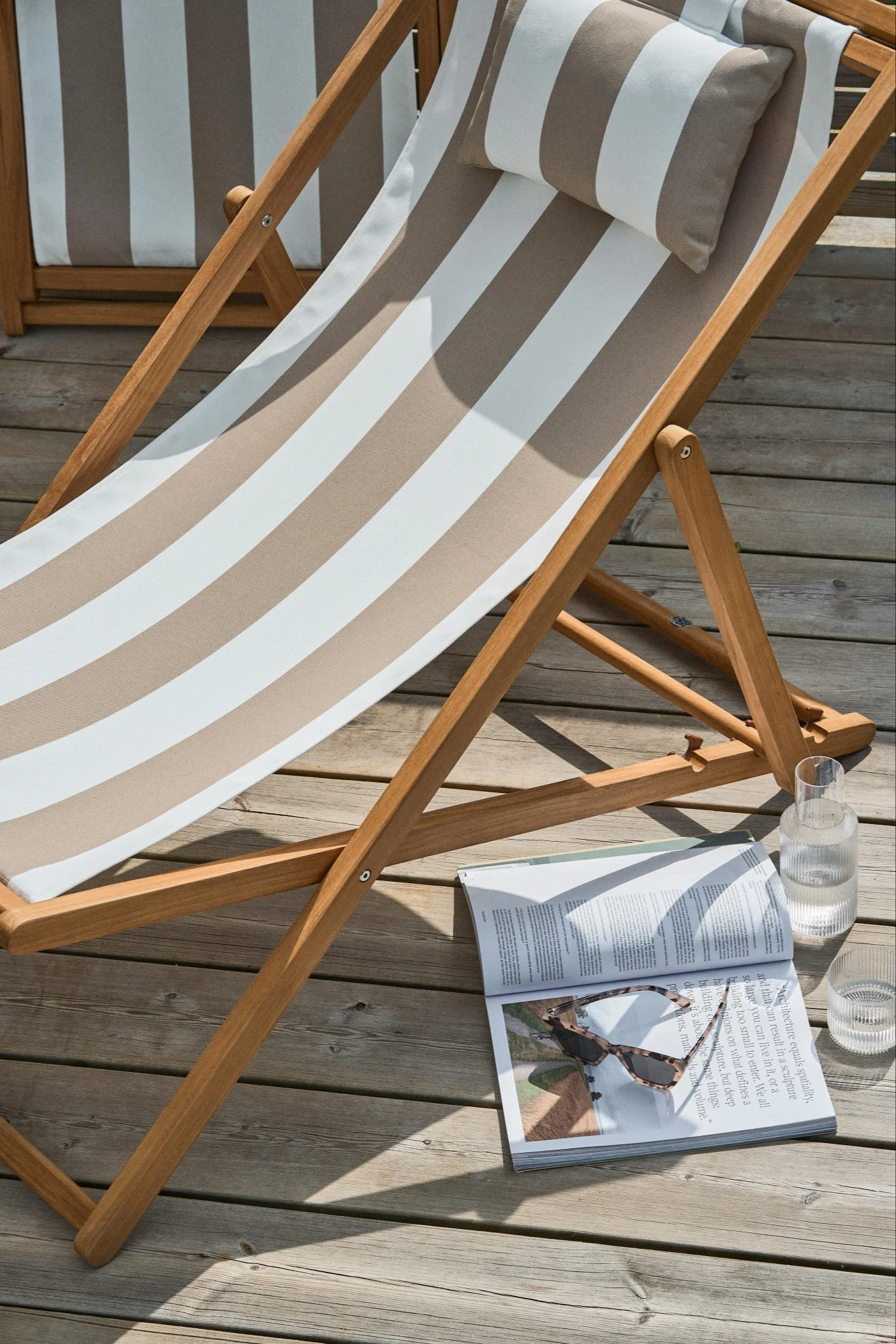 A wooden lounge chair with a striped cushion and pillow, a magazine with sunglasses, and a glass of water on a wooden deck.