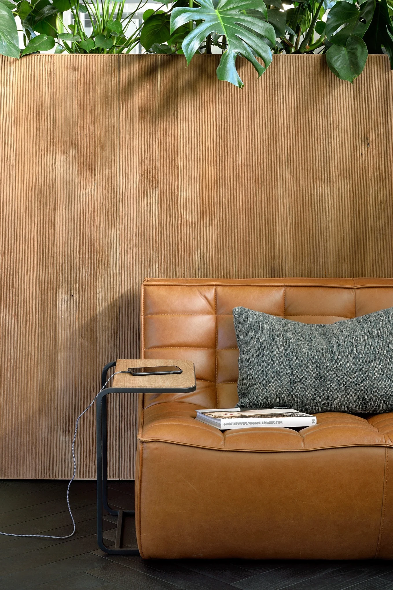 A tan leather sofa with a textured grey pillow next to a small side table with a charging phone, in front of a fluted wood wall with plants.