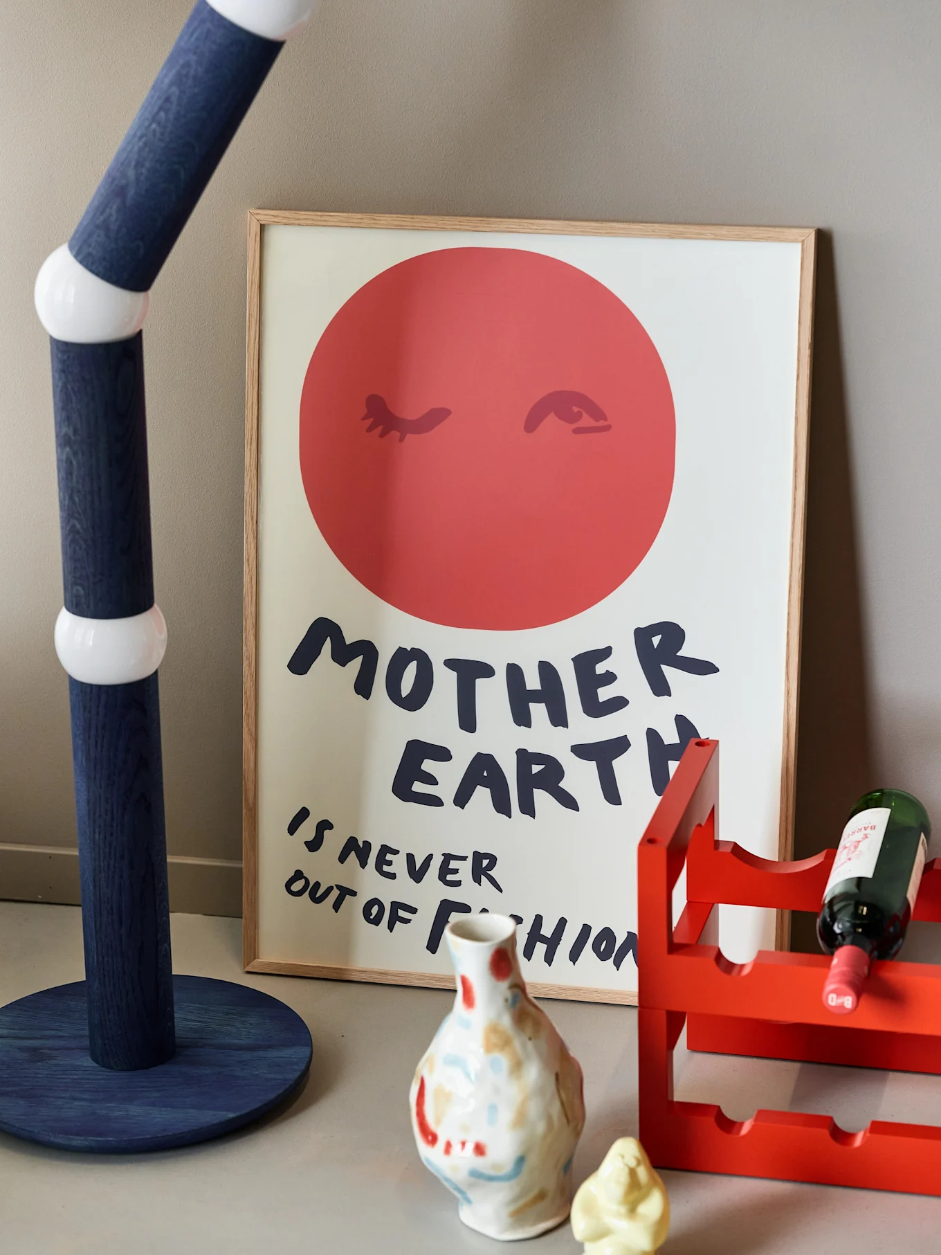 A framed poster with "MOTHER EARTH IS NEVER OUT OF FASHION" text, a blue floor lamp, and a red wine rack with a bottle.