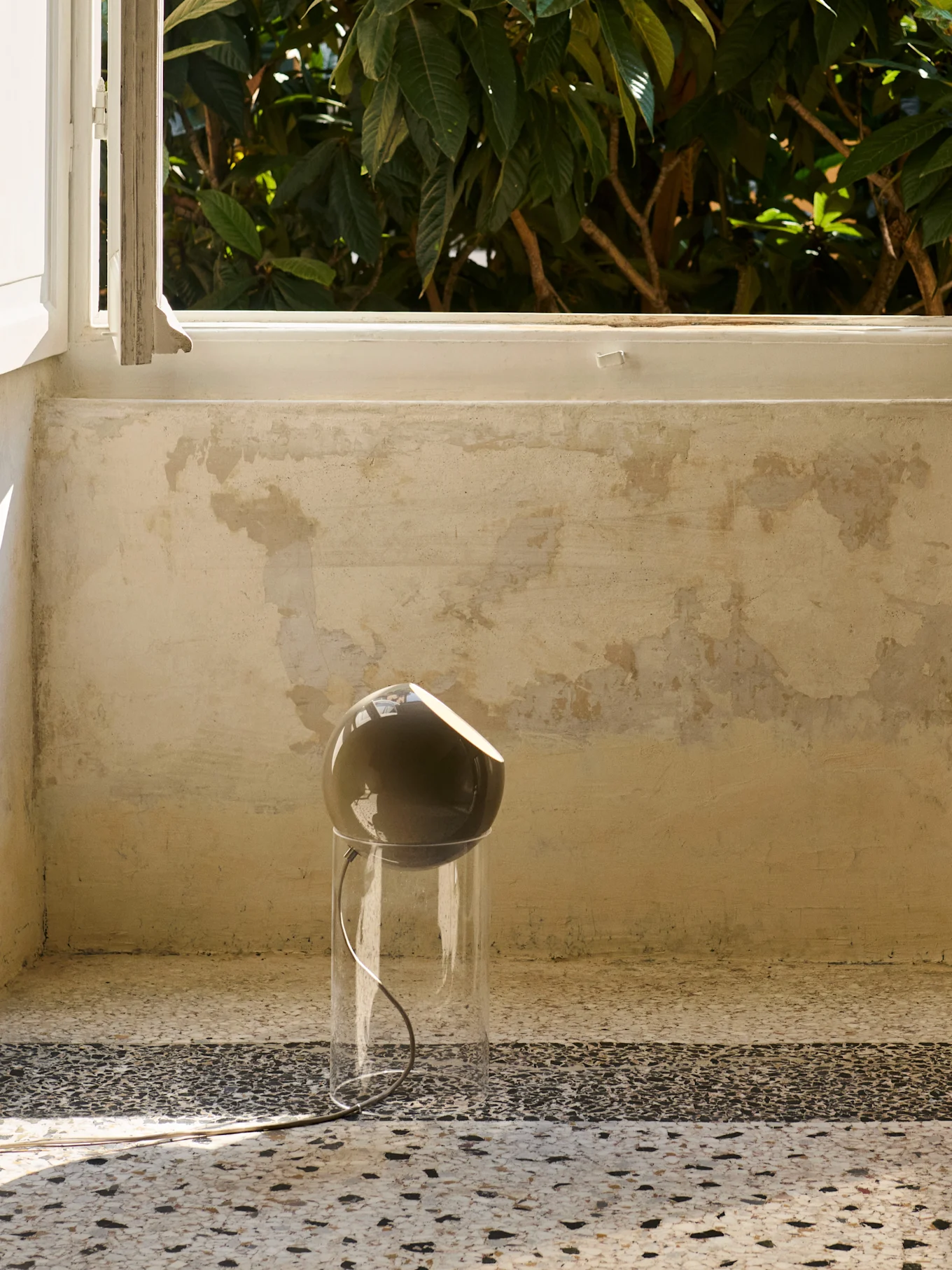 A modern lamp with a dark, spherical head and a clear cylindrical base sits on a speckled floor by a window with green foliage outside.