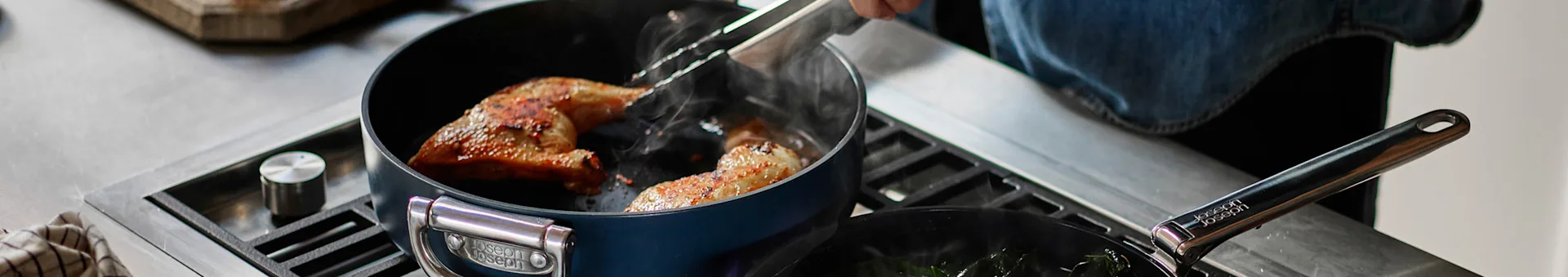 Person using tongs to flip chicken in a blue pot on a gas stove, with another pan of greens to the side.