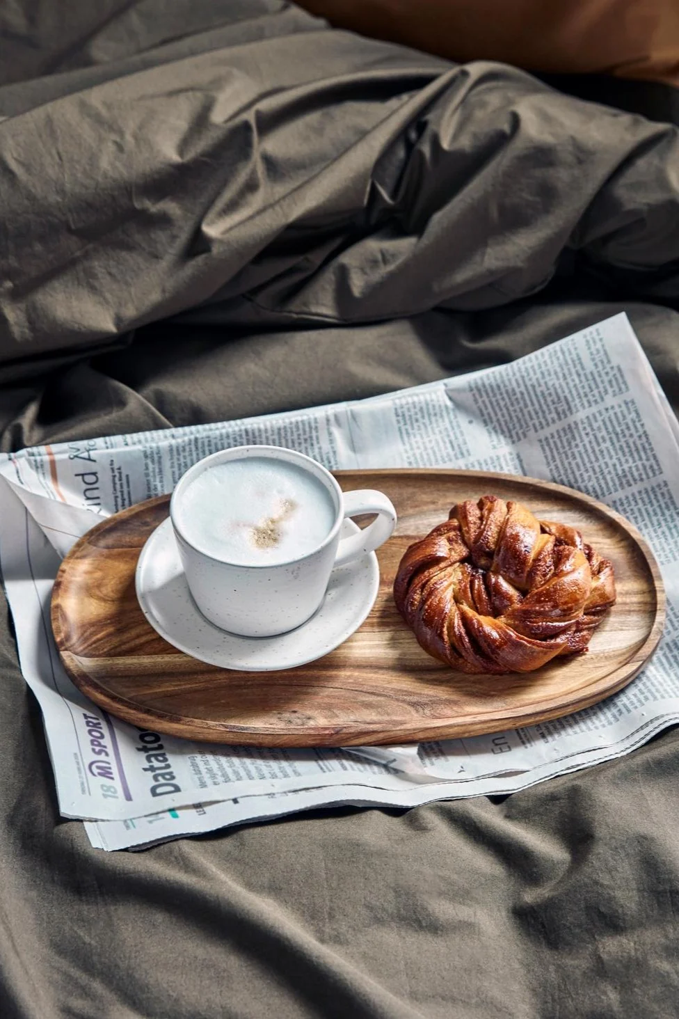 Coffee and a pastry on a wooden tray with a newspaper, set on dark bedding.