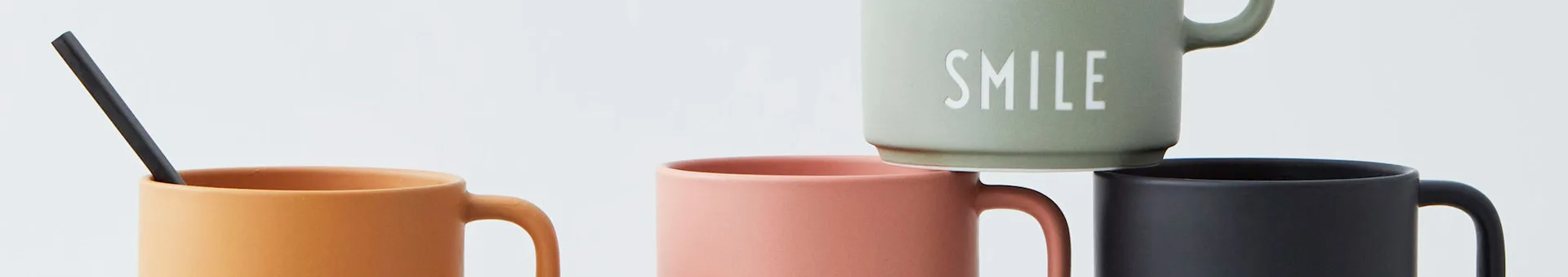 Four ceramic mugs, an orange one with a black straw, a pink one, a black one, and a green 'SMILE' mug stacked on top.