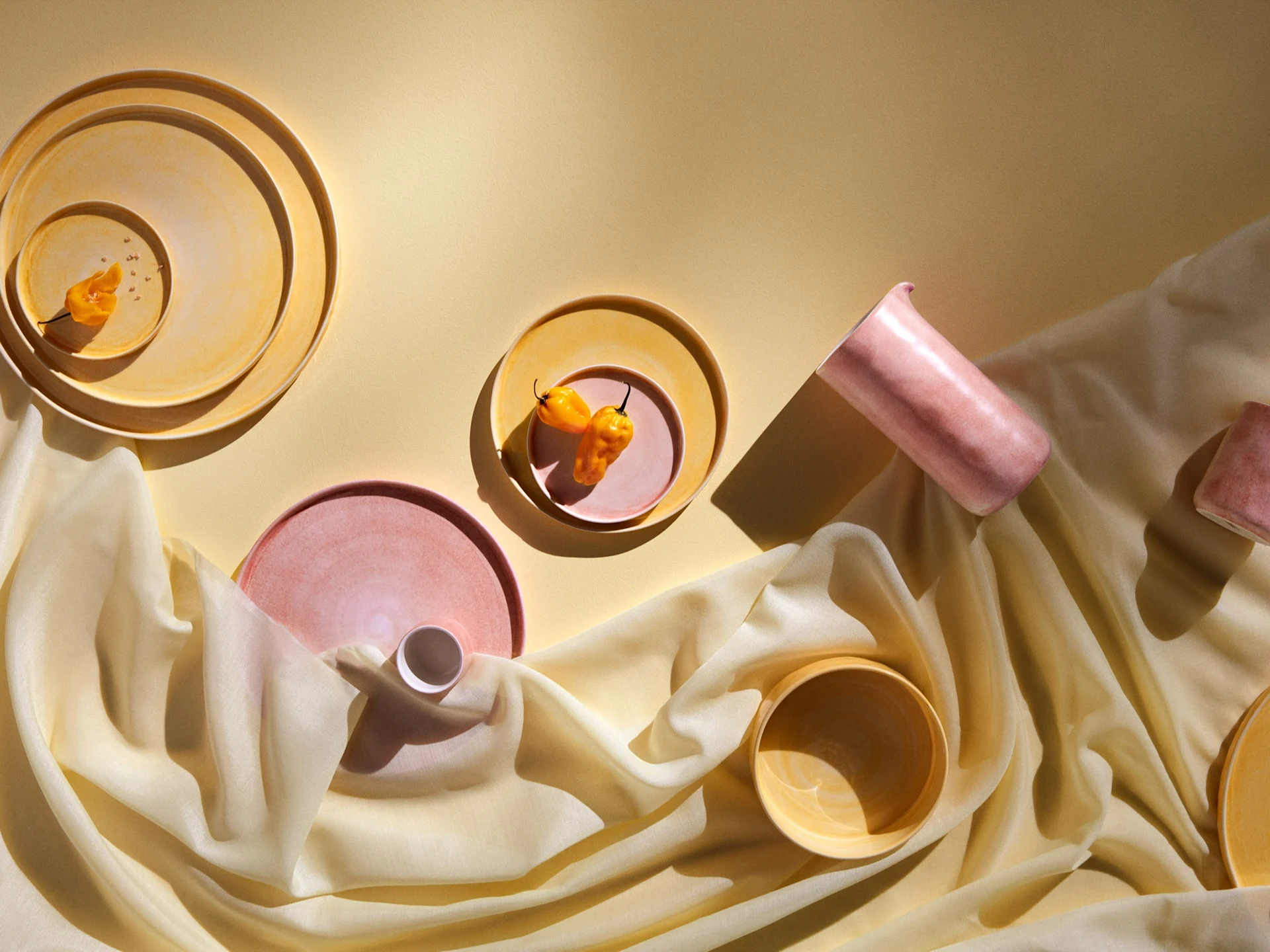 Yellow and pink ceramic plates, bowls, cups, and a pitcher on draped fabric and a yellow surface, with yellow peppers.