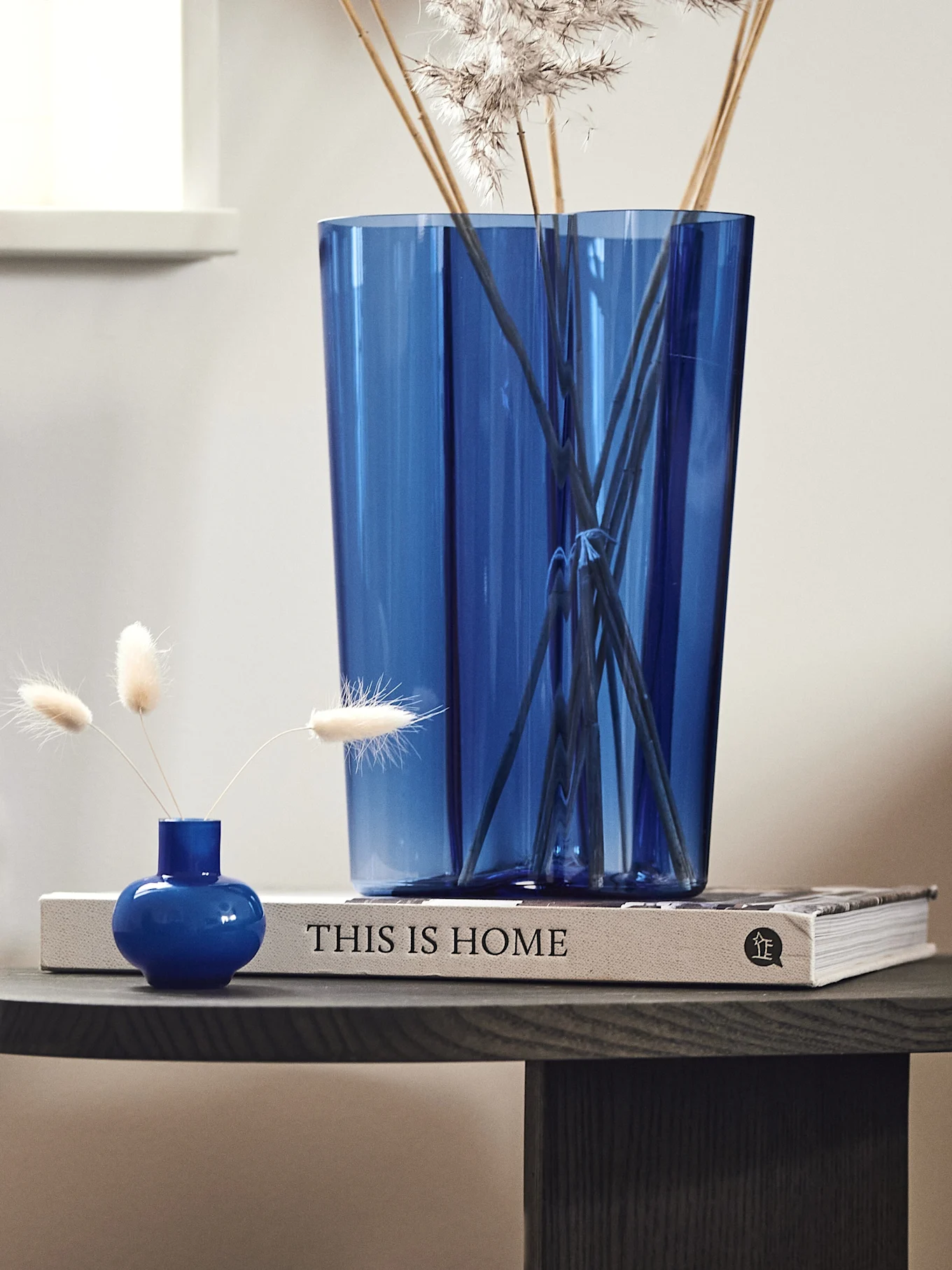 Two blue vases, one large with dried grasses and one small with three fluffy dried flowers, on a book titled 'THIS IS HOME'.