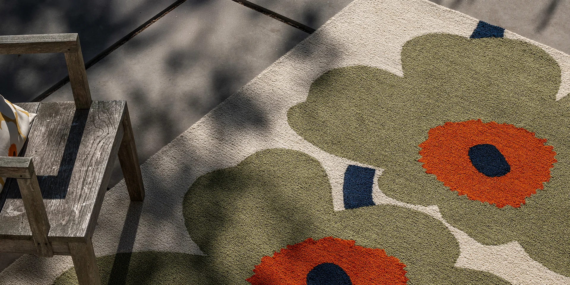 Olive green and orange floral outdoor rug next to a weathered wooden chair on a concrete patio.