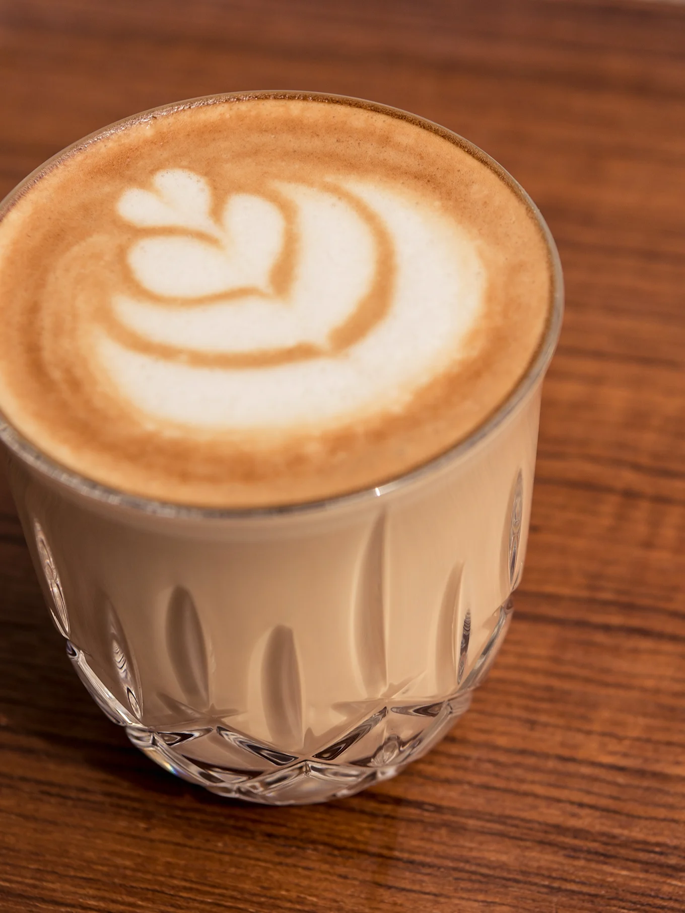 Close-up of a latte with heart-shaped art in a clear faceted glass on a wooden table.