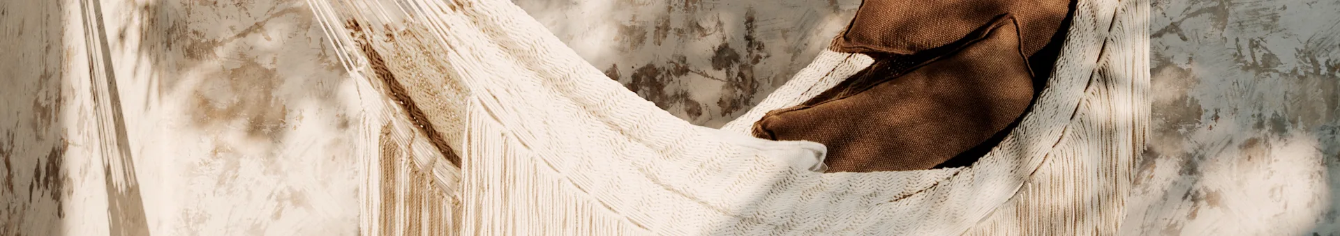 Close-up of a cream-colored woven hammock with fringes and a brown textile resting in it, against a textured wall with shadows.