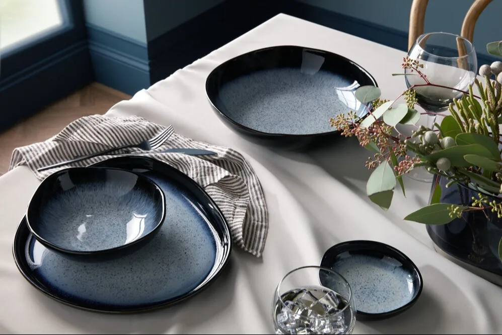 Blue and black speckled dinnerware set on a white tablecloth, featuring plates, bowls, and a large serving dish, with cutlery and a floral centerpiece.