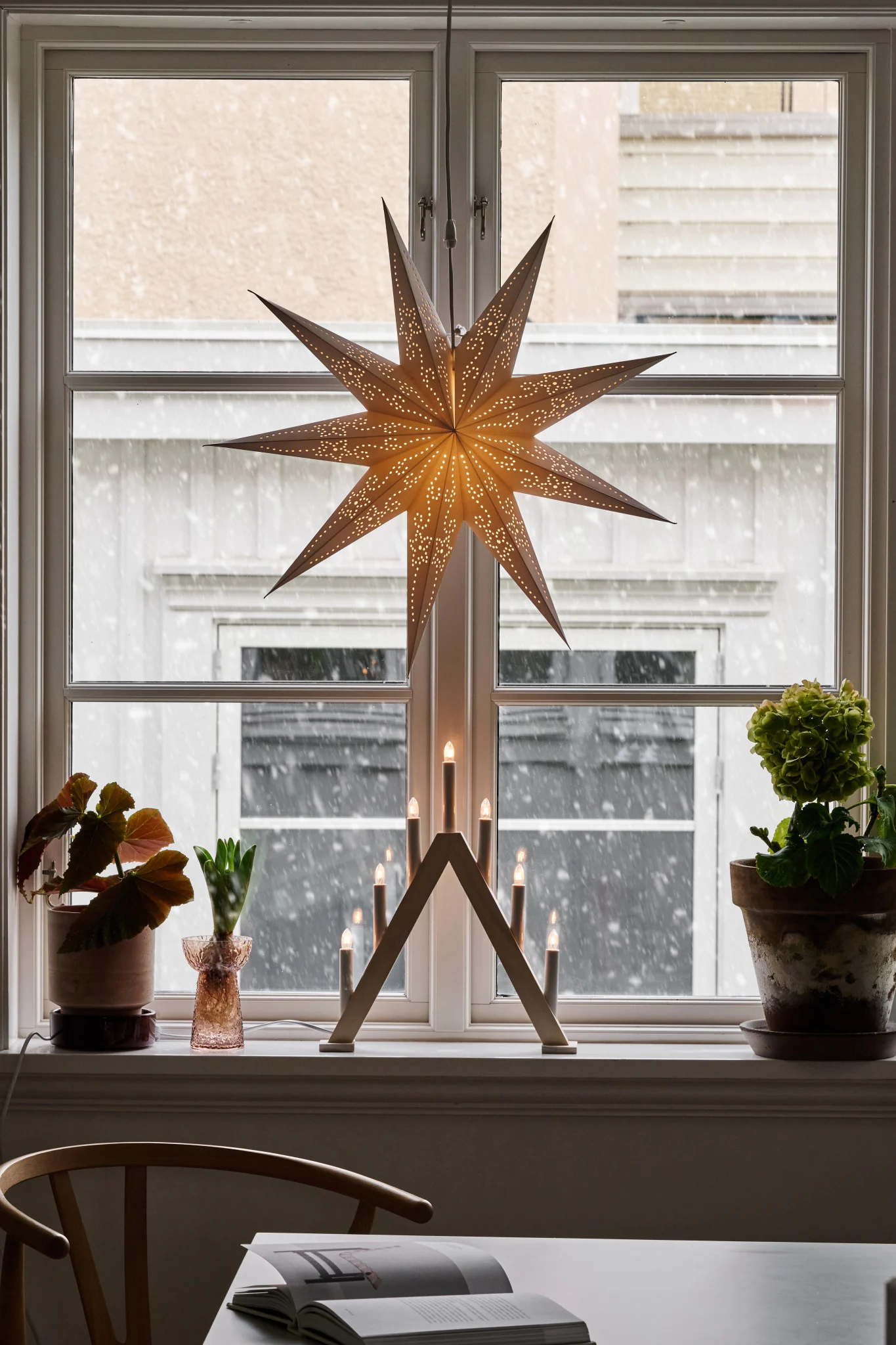 A glowing paper star and a wooden candelabra on a window sill, with snow falling outside.
