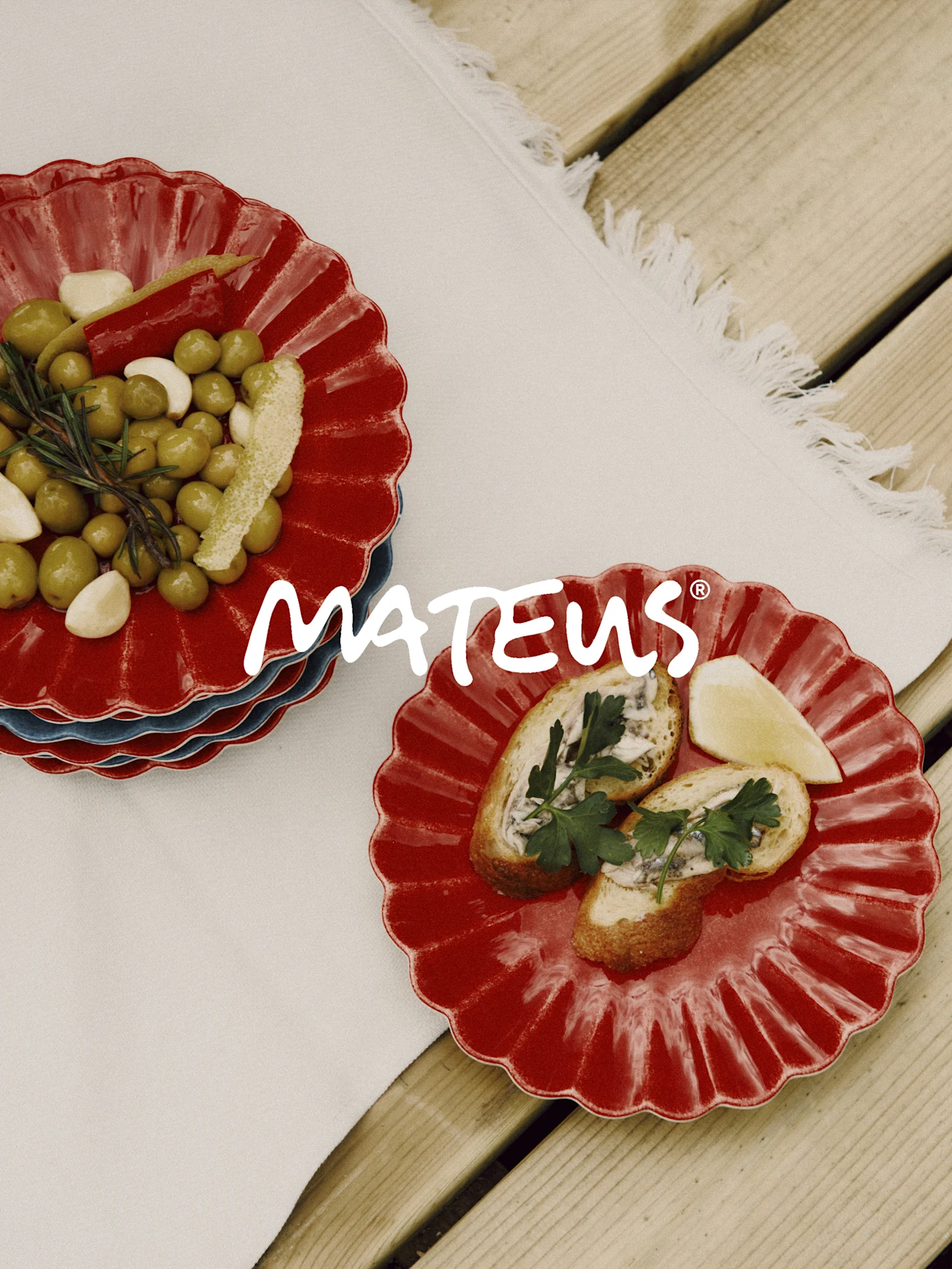 Red scalloped plates with olives, garlic, rosemary, and bread with anchovies on a wooden table. MATEUS brand.