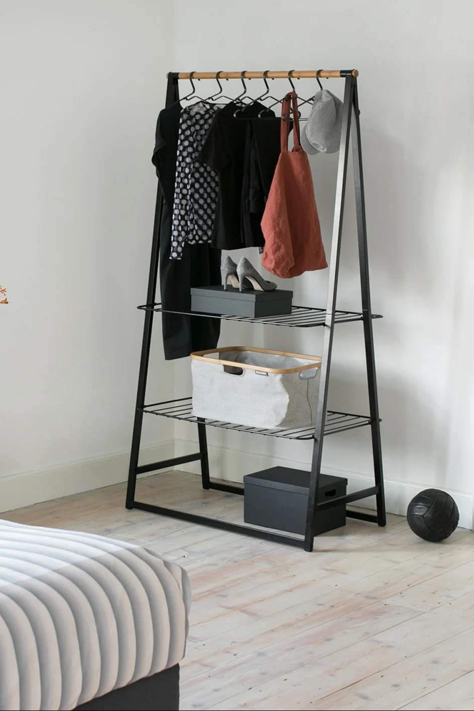 Modern black A-frame clothes rack with wooden rail, clothes, shoes, basket, and storage boxes on light wood floor.