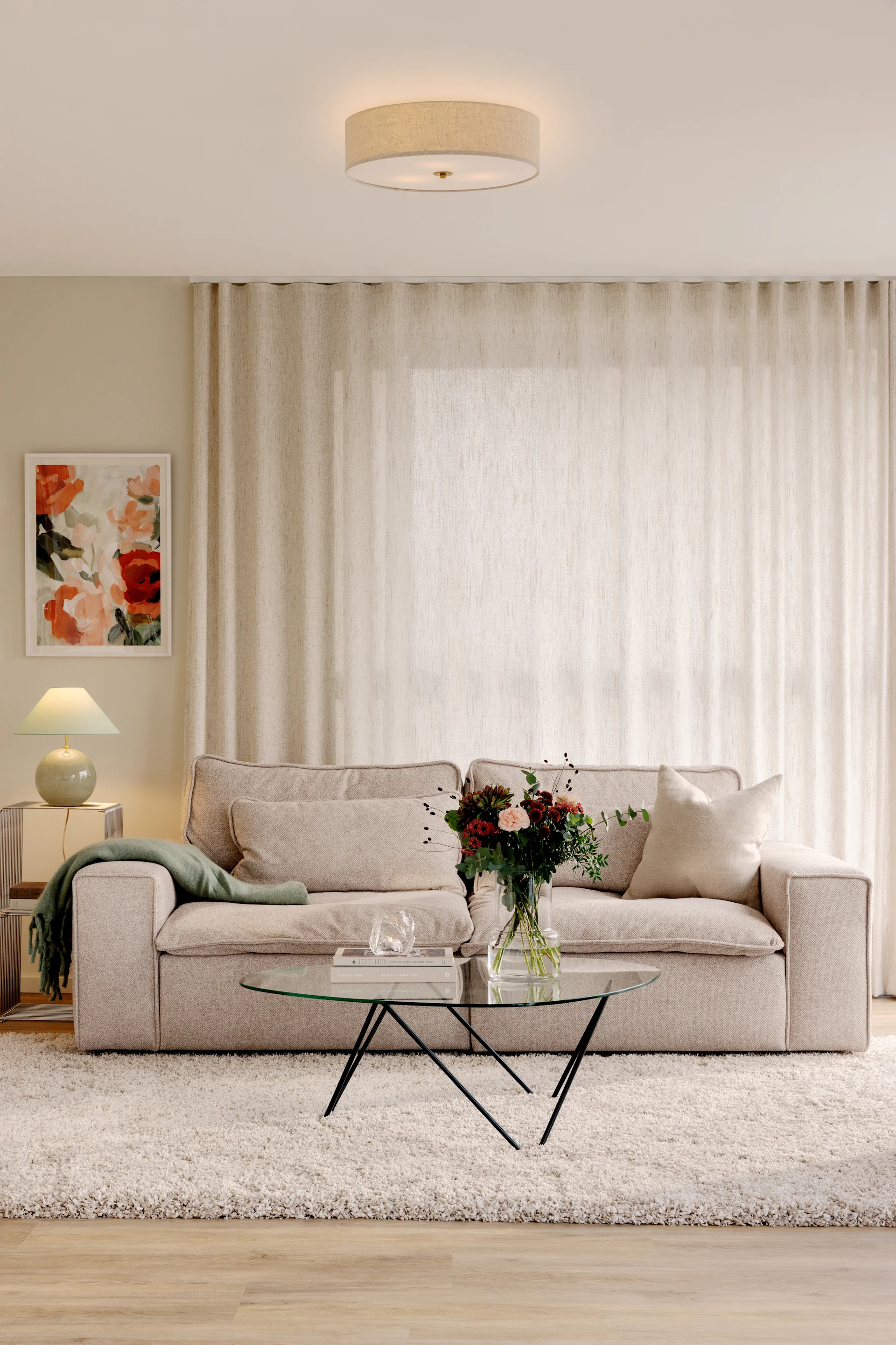 Cozy living room with a light fabric sofa, glass coffee table with flowers, side lamp, and abstract floral art.