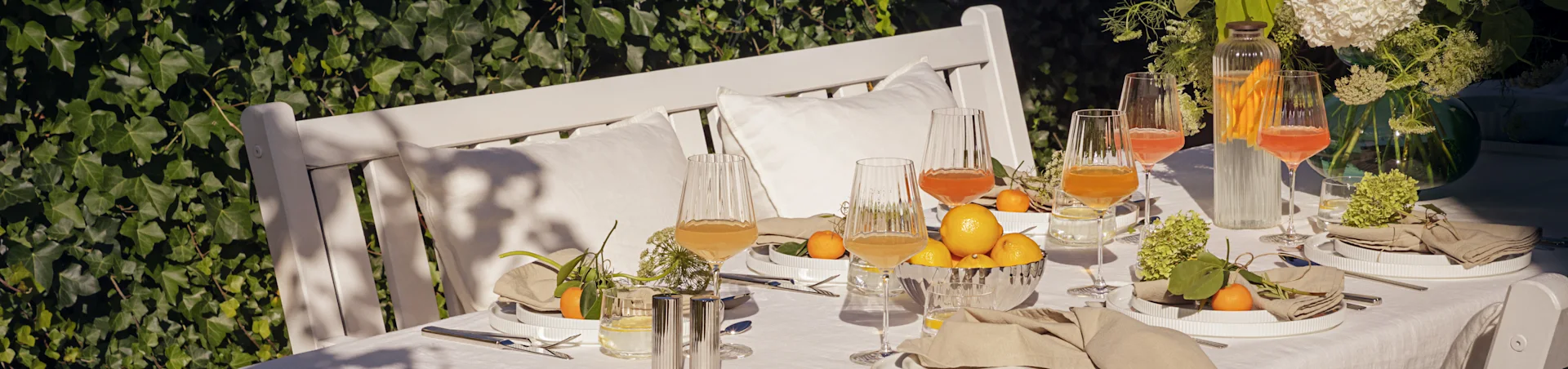 Outdoor table set for a summer meal with white ridged plates, linen napkins, fluted glasses with drinks, lemons, and mandarins.