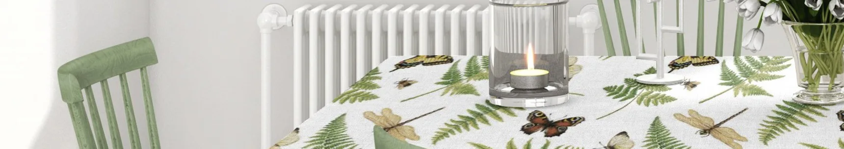 White tablecloth with green fern, dragonfly, and butterfly pattern, holding a lit candle in a glass holder and a vase of white tulips.