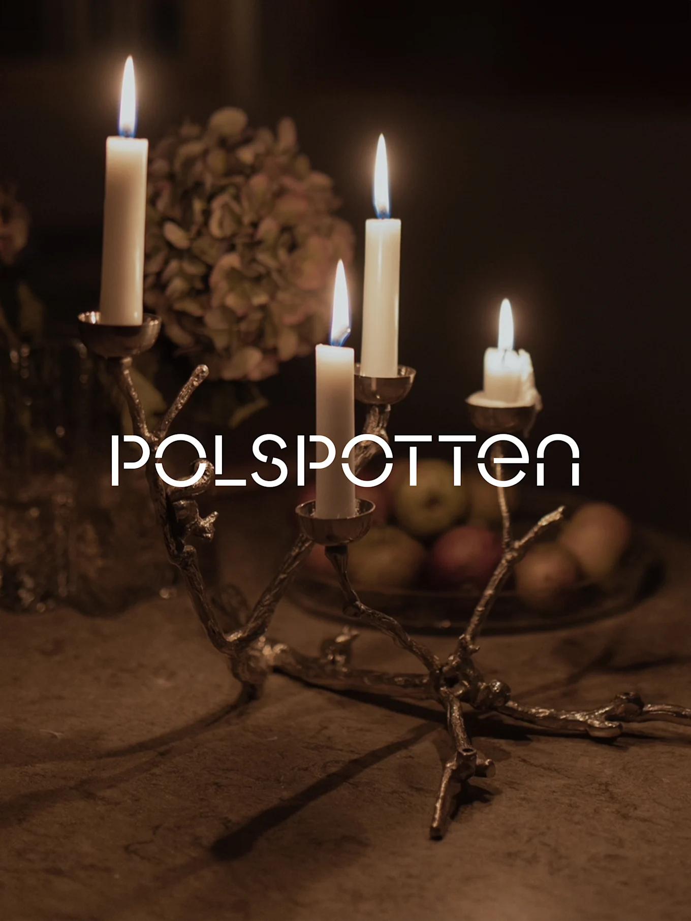 Golden, branch-like candelabra with four lit white candles, against a dark, warm background with blurred flowers and fruit.