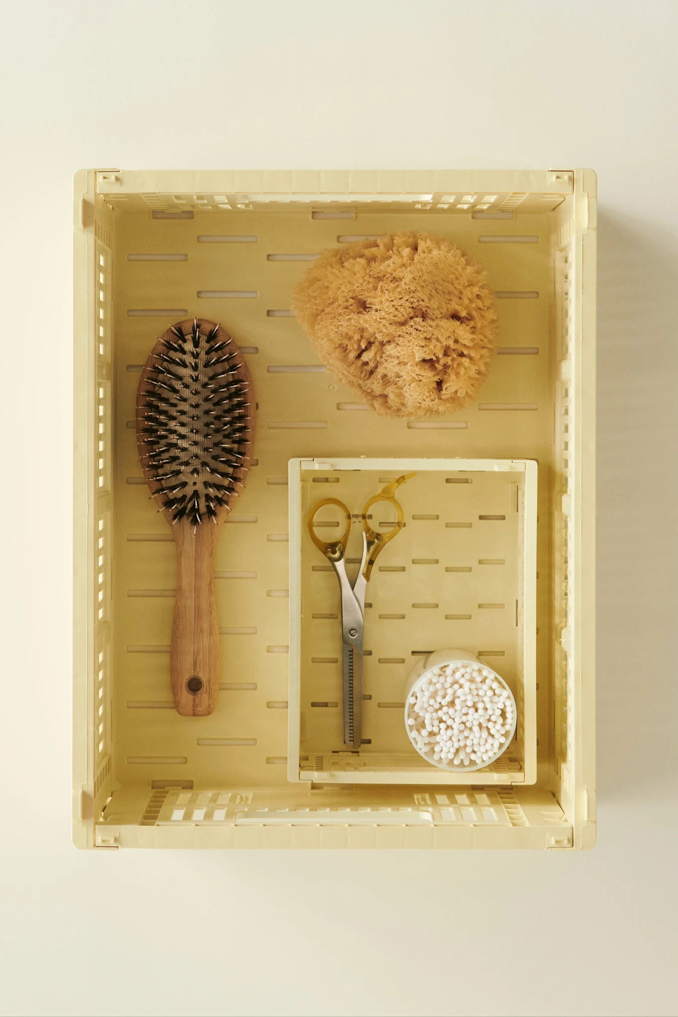 Light yellow crate holds a wooden hairbrush, natural sponge, hair shears, and cotton swabs, all on a light beige background.