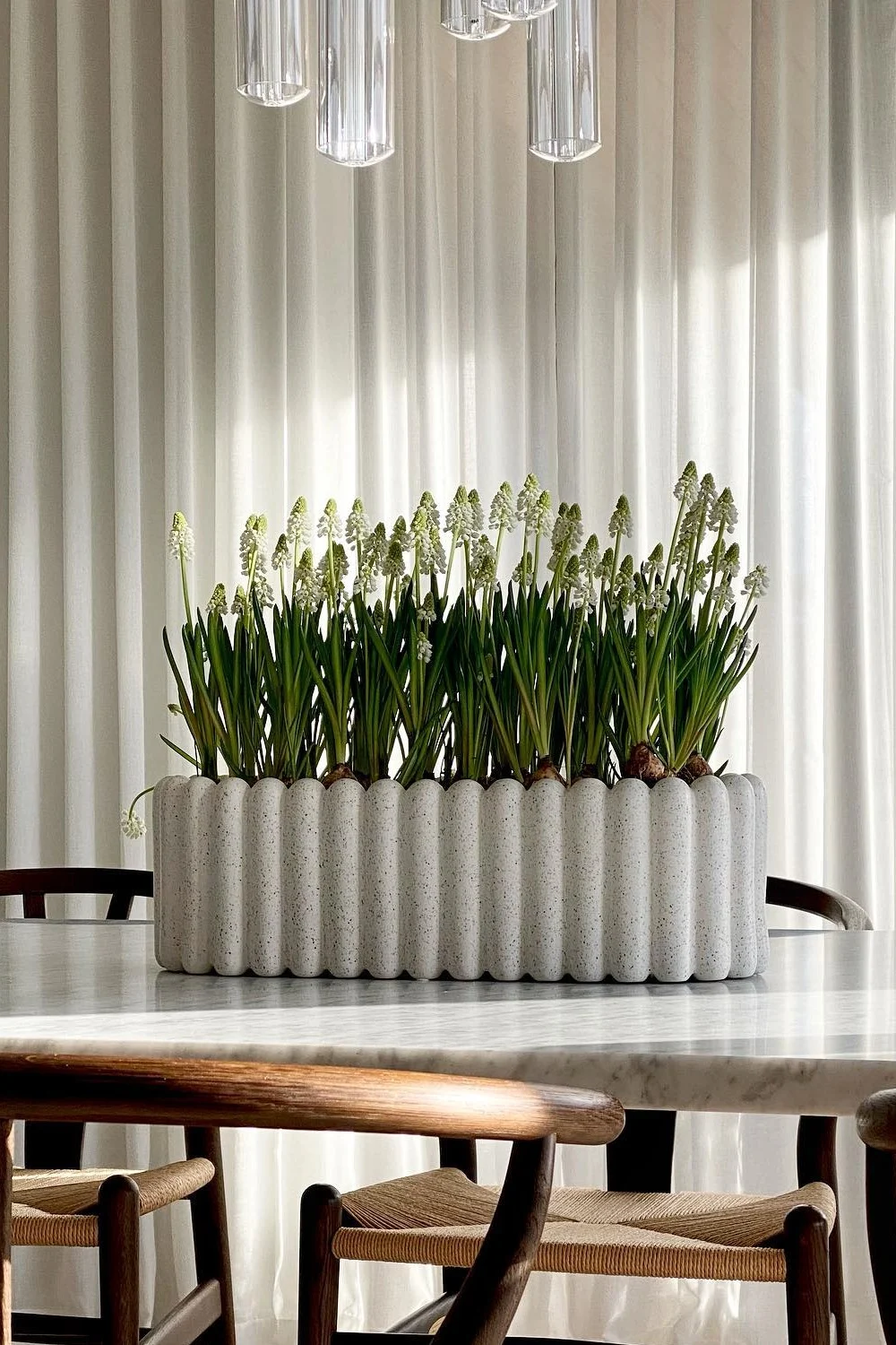 A long, white speckled wavy planter filled with small white bell-shaped flowers sits on a marble table.