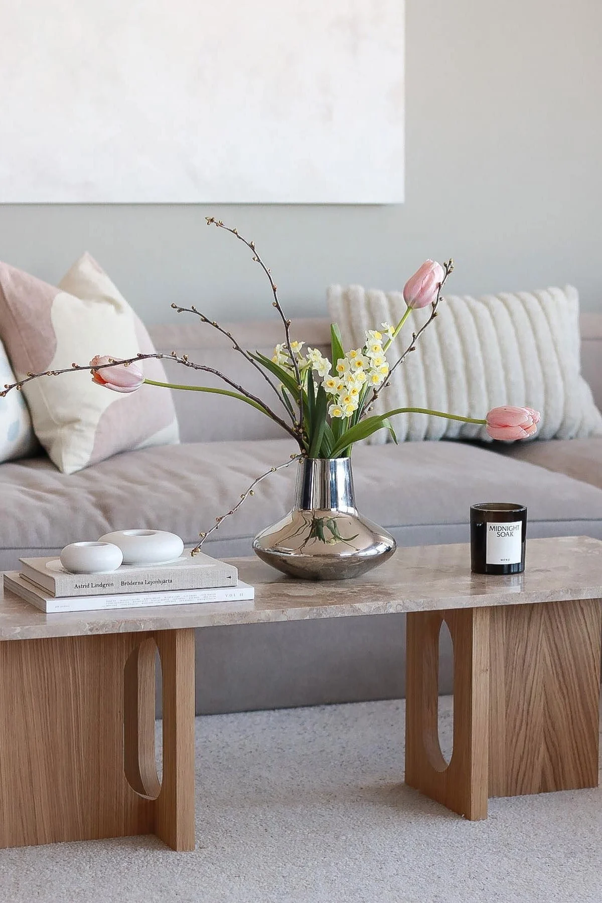 A silver vase with pink tulips, white narcissus, and branches sits on a marble coffee table, with books, white decor, and a dark jar candle.