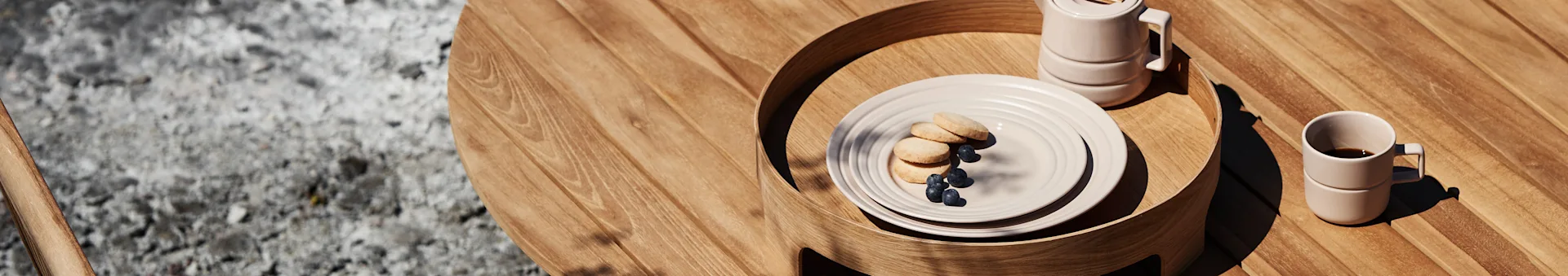 Outdoor wooden table with a light tray holding plates of cookies, blueberries, a pitcher, and stacked coffee mugs.