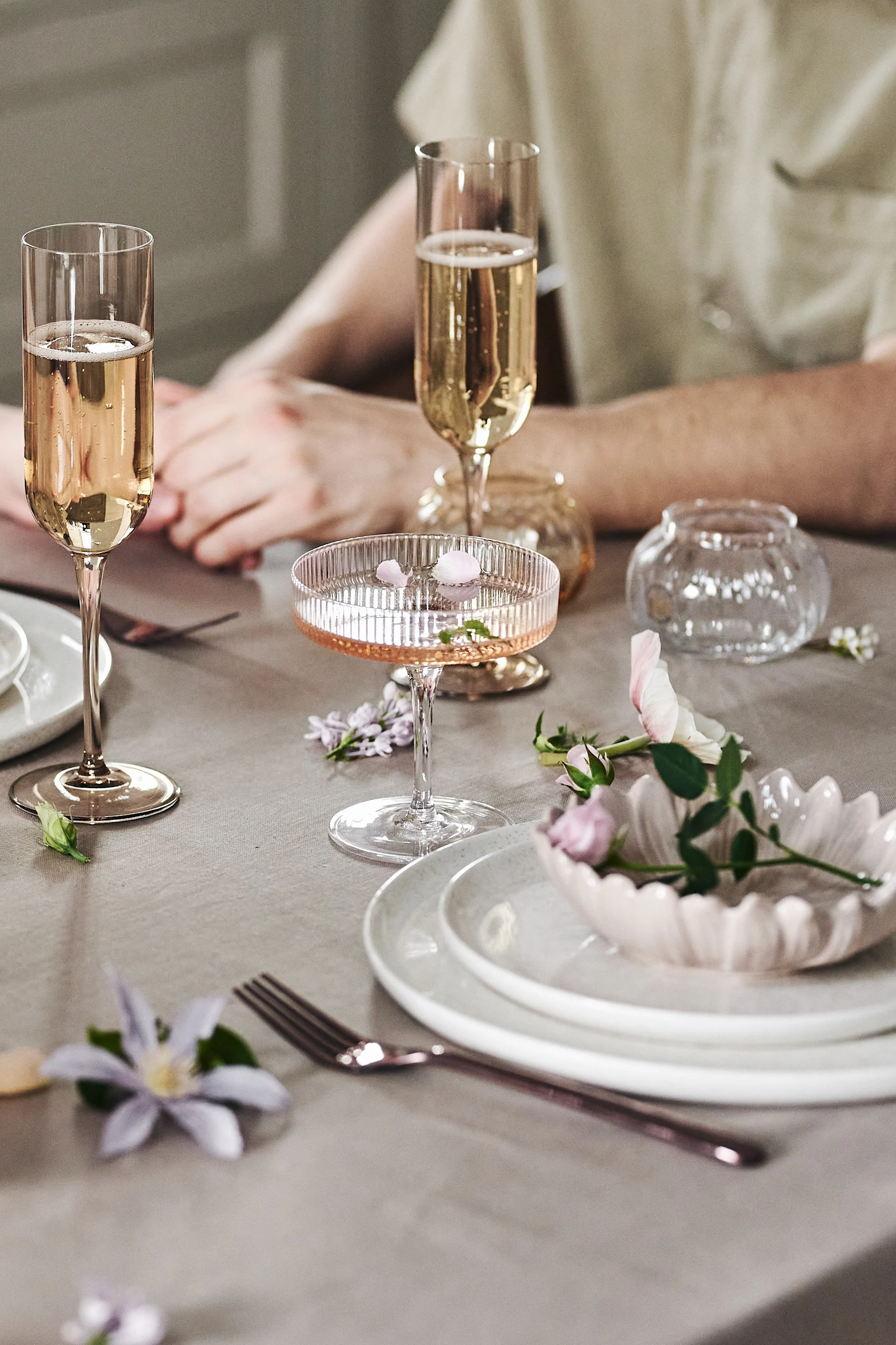 Elegant table set with two flutes and one coupe glass of sparkling wine, plates, a fork, and scattered pink and purple flowers.