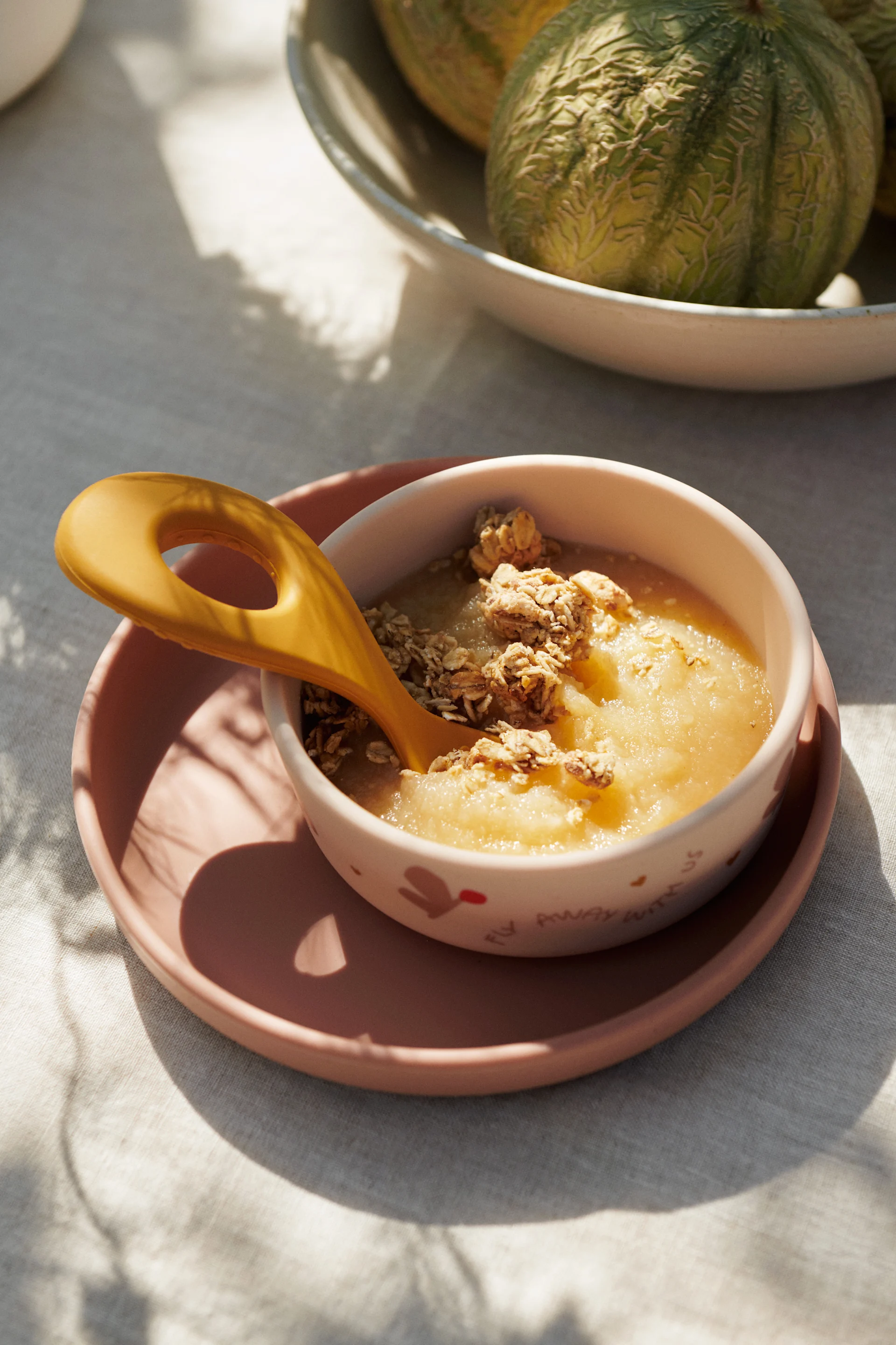 Applesauce with granola and a yellow spoon in a bowl on a pink plate, with melons.