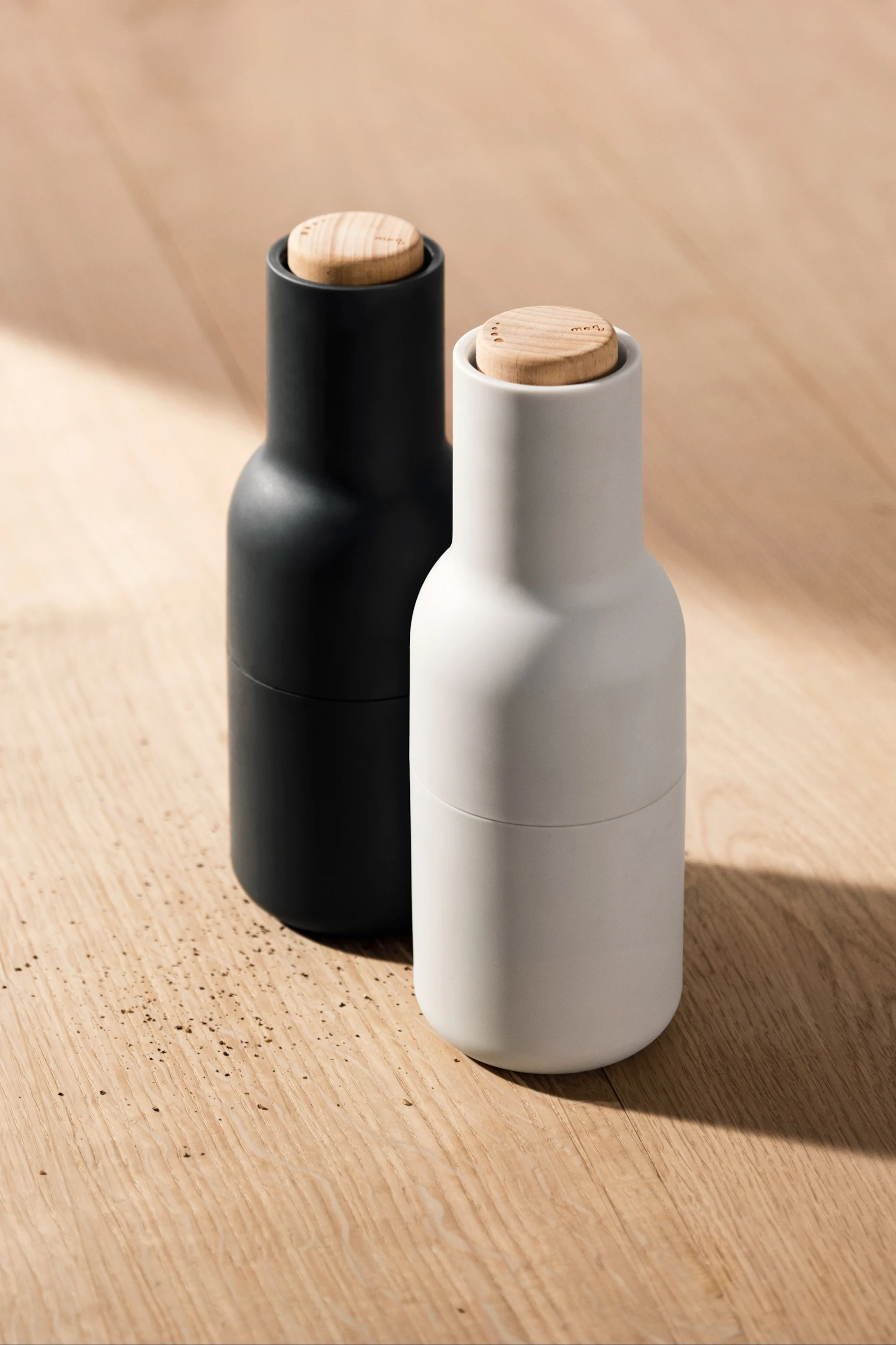 Two minimalist spice grinders, one black and one white, with light wooden caps, on a wooden surface.