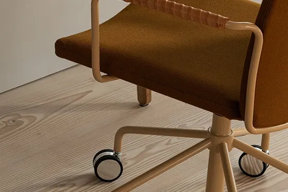 A close-up of an office chair with a brown fabric seat, light beige metal frame, and casters, on a light wooden floor.