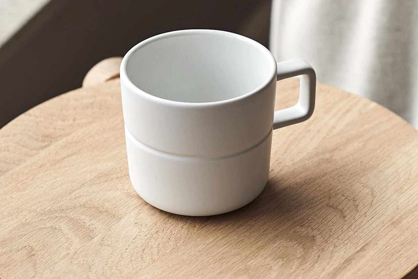 A white ceramic mug with a handle sitting on a light wooden surface. The mug has a subtle horizontal band around it.