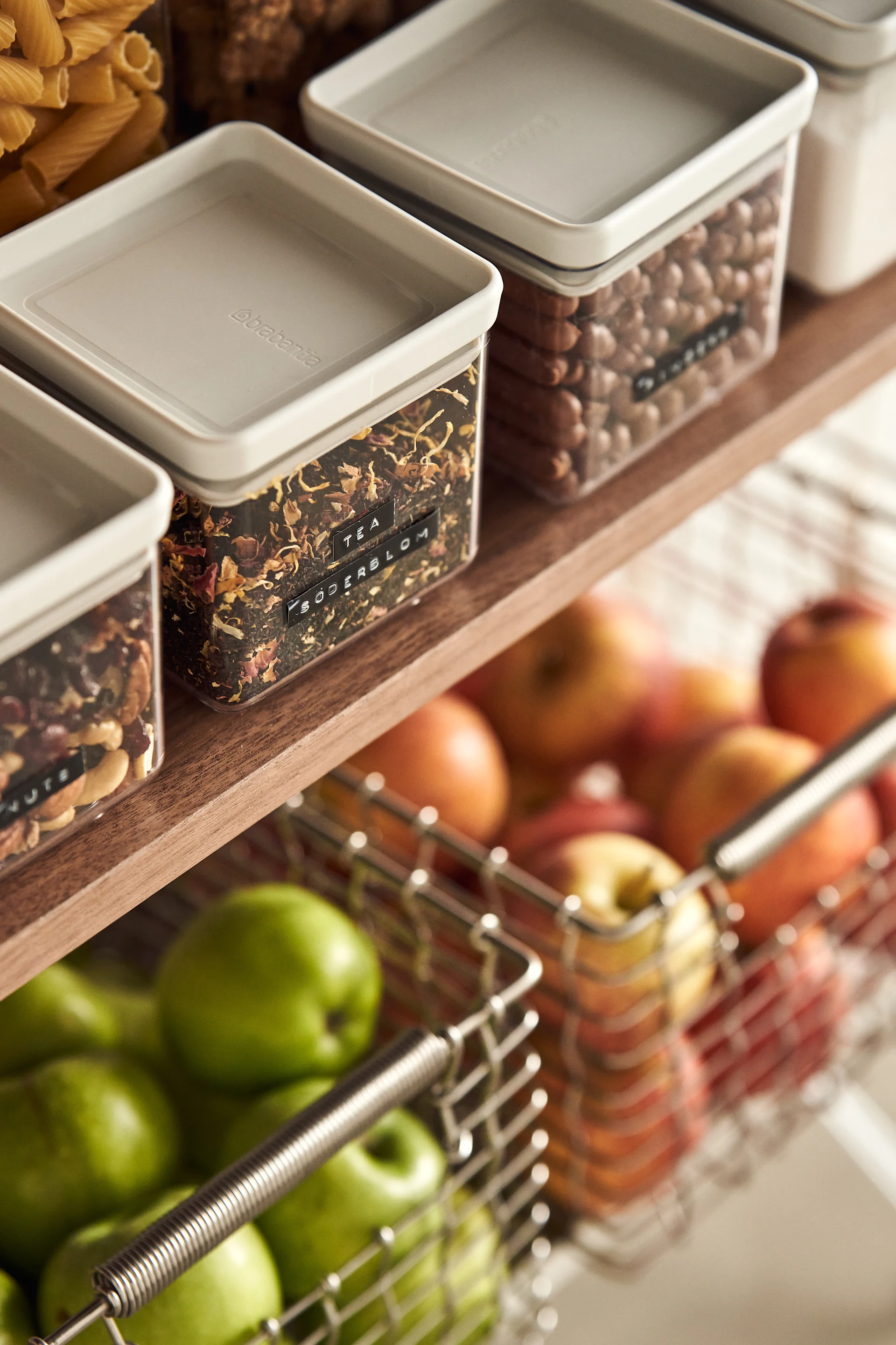 Transparent containers with light grey lids filled with tea, nuts, and pasta on a wooden shelf, above wire baskets of green and red apples.