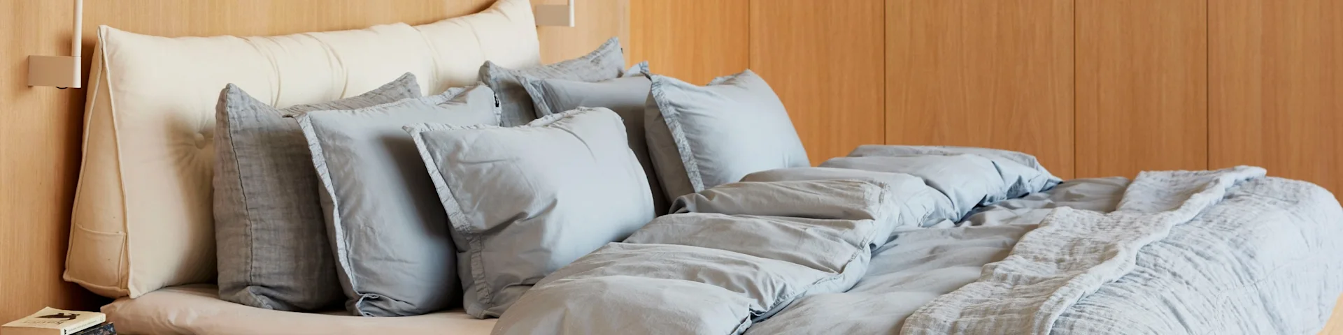 Bed with layered bedding: an off-white pillow, several light gray pillows, a gray duvet, and a textured light gray throw blanket. A light wood paneled wall is behind.