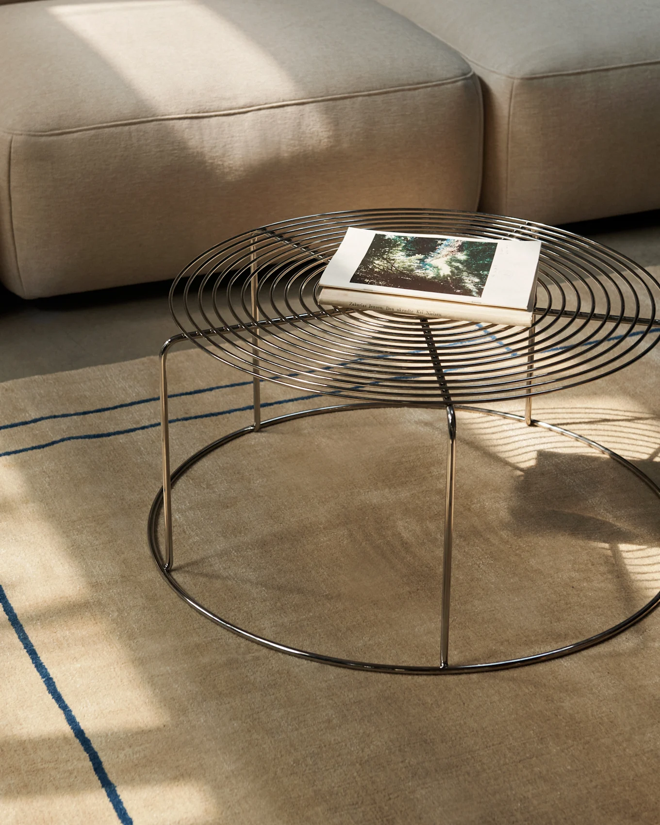 Round metal coffee table with a book on a beige rug, next to a sofa.