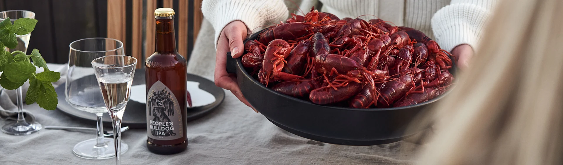 Person holding a black bowl filled with cooked red crayfish on a linen-covered table with a beer bottle and glasses.