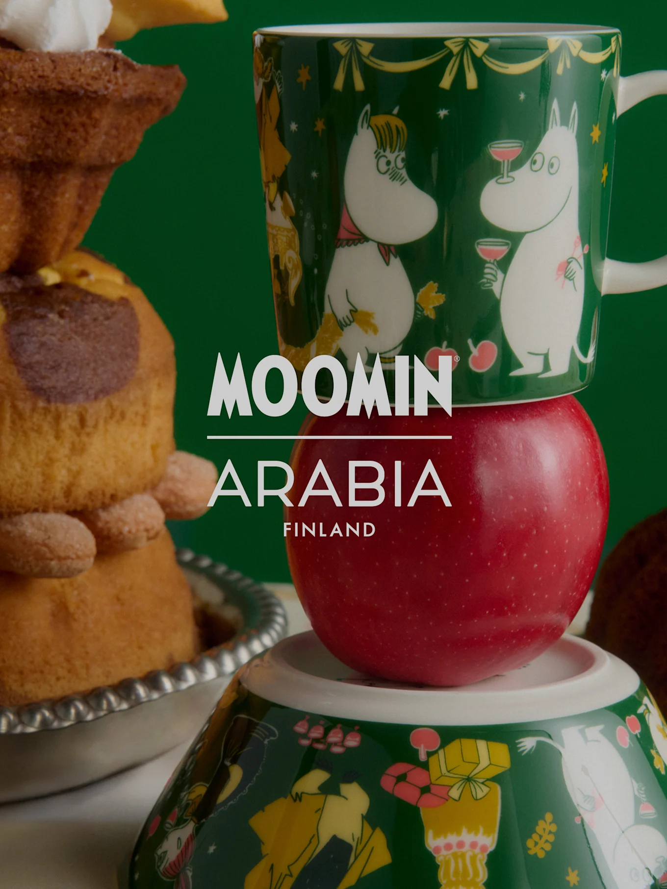 Moomin Arabia Christmas collection: A green Moomin mug on a red apple, atop a Moomin bowl, beside festive baked goods.